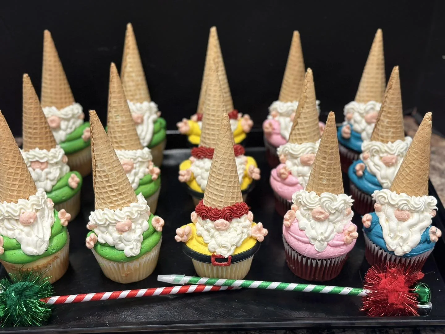 Cupcakes decorated as gnomes wearing colorful outfits and cone hats, arranged on a black tray with holiday decorations.