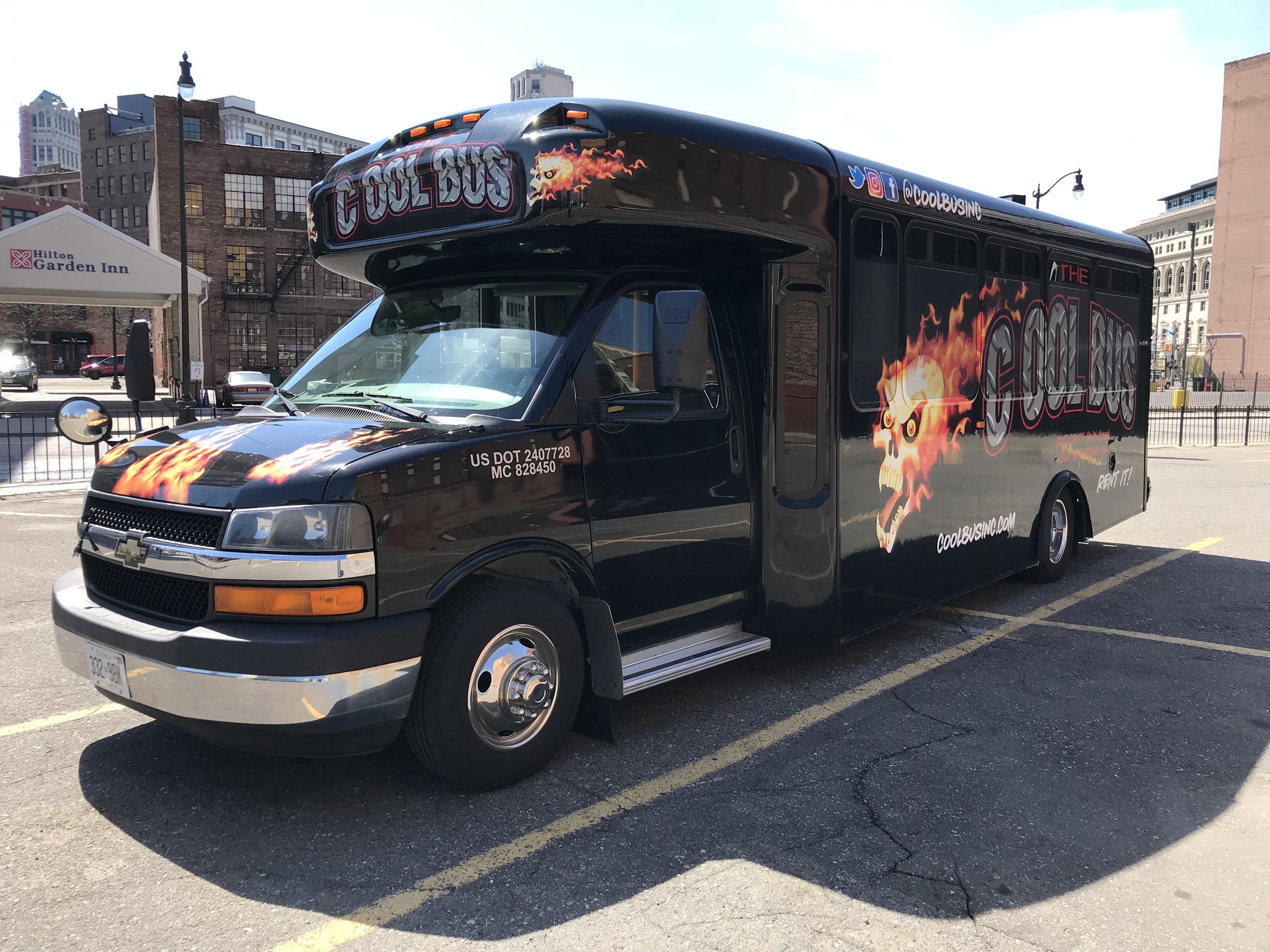 A black party bus named "The Cool Bus" with flame graphics and skull designs, parked in a lot near a Hilton Garden Inn.