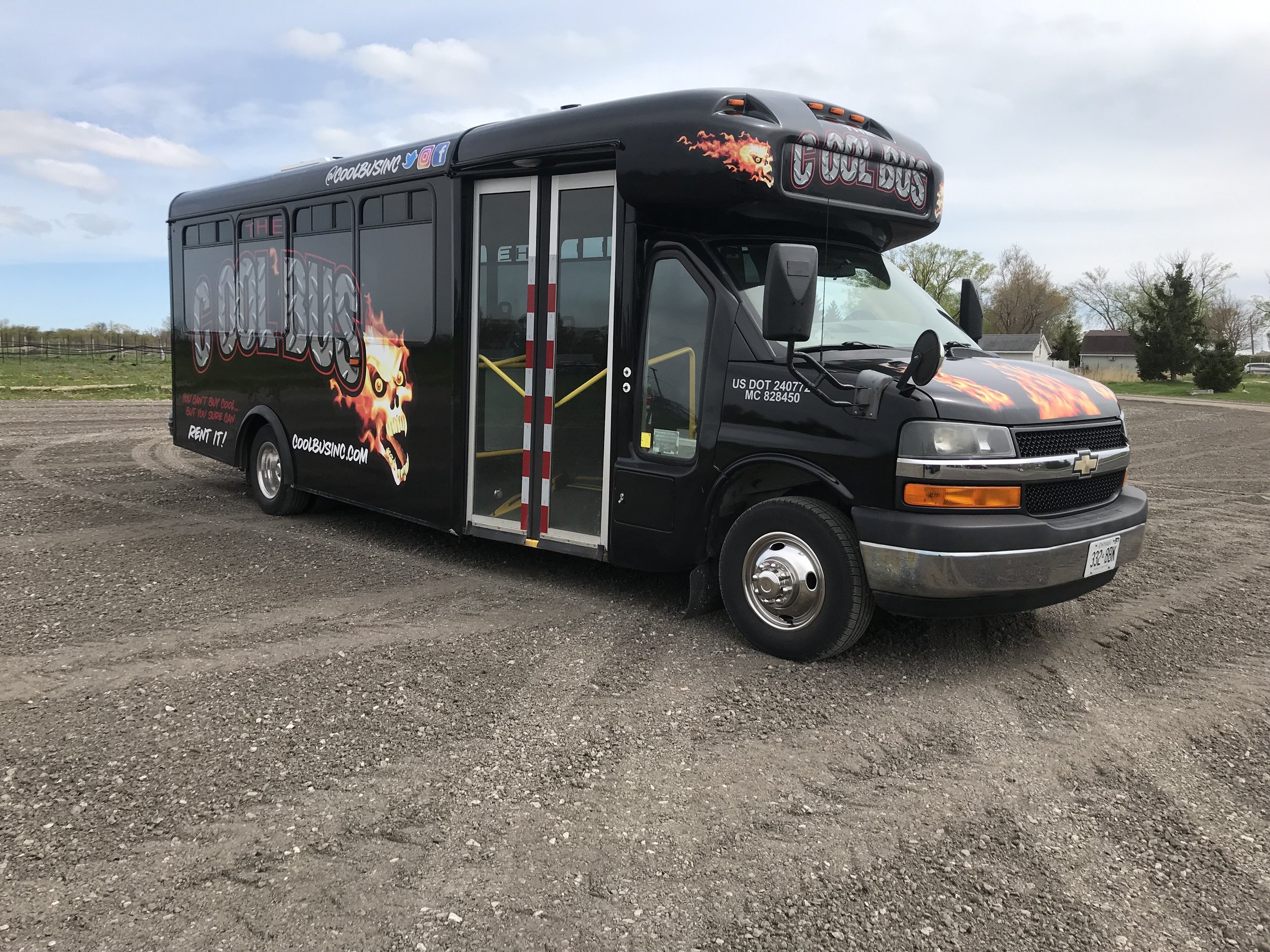 Black party bus with 'The Cool Bus' branding, flame designs, parked on gravel.