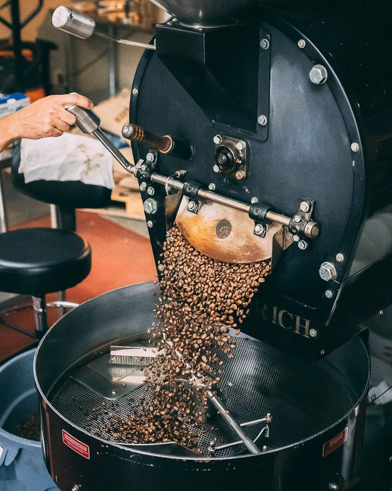 A person operating a coffee roaster with coffee beans inside, pouring roasted beans into a metal container.