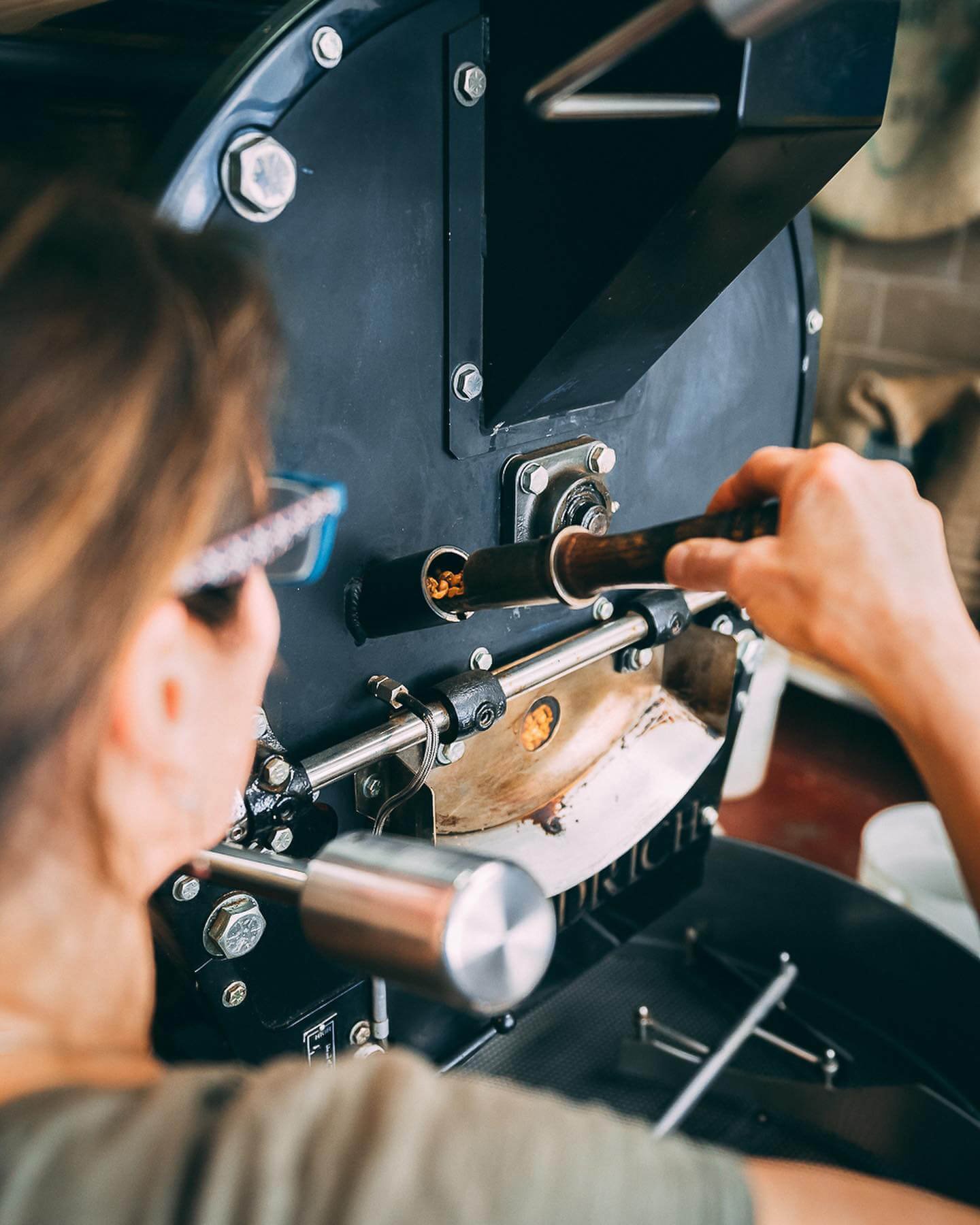 A person wearing glasses operates a vintage coffee roaster machine, inserting green coffee beans into the roasting chamber with a handle, with the machine's metallic parts and knobs visible.