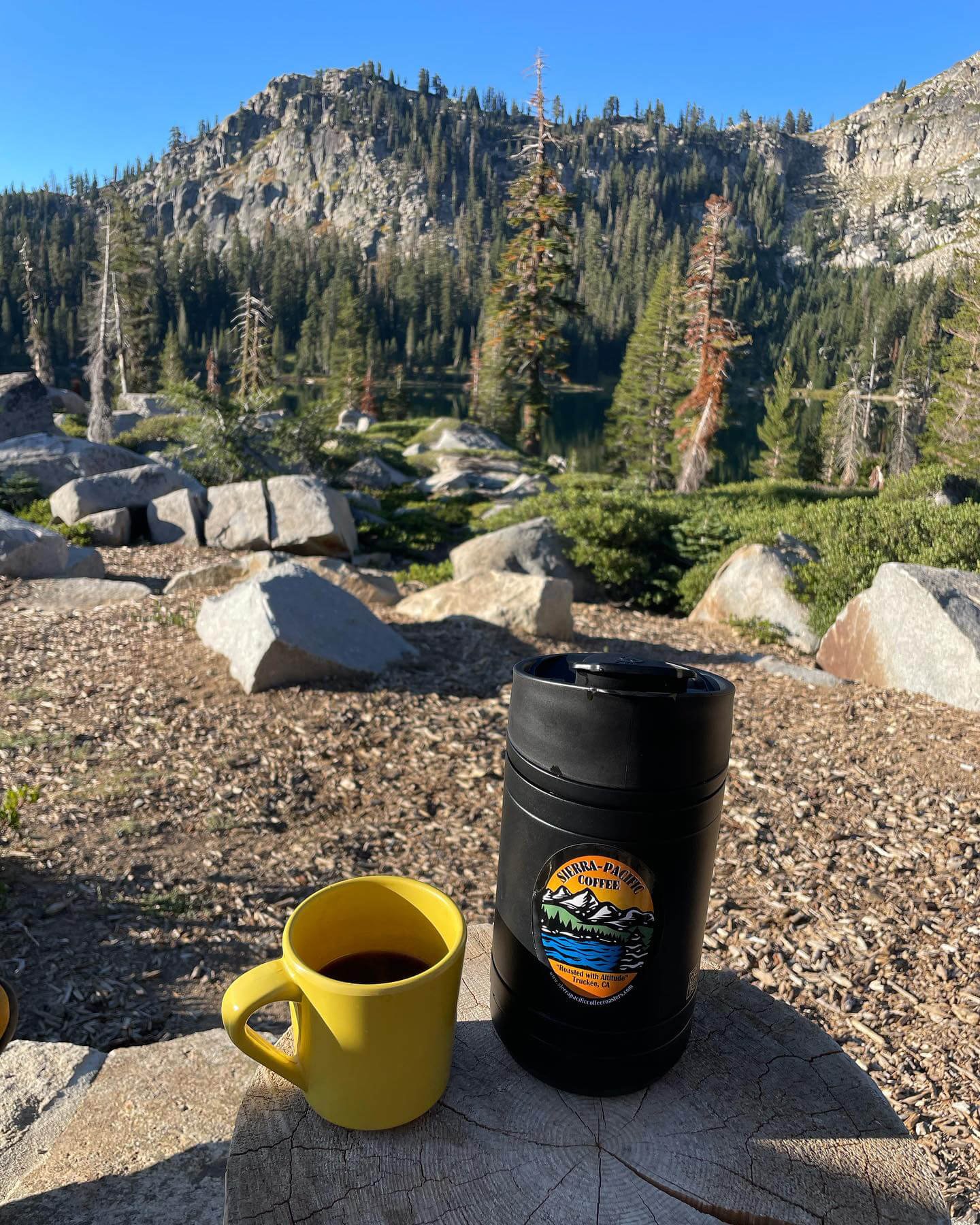 A yellow mug and a black thermos with a Sierra Pacific Coffee logo sit on a wooden log table outdoors in a mountainous forest. Tall pine trees and rocky terrain are visible in the background under a clear blue sky.