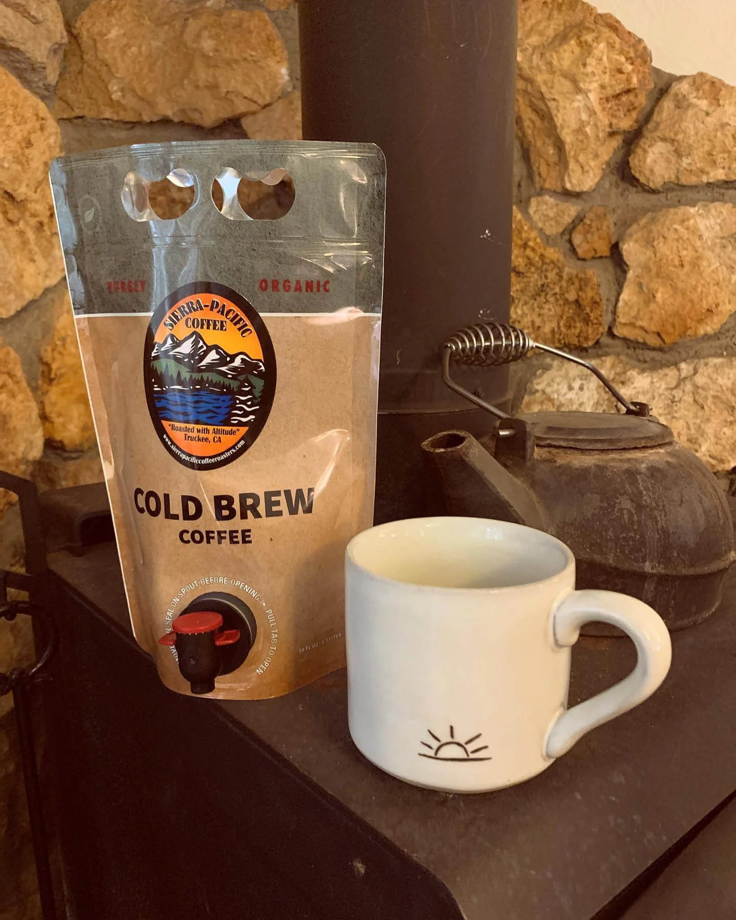A bag of Ultra Pacific Cold Brew Coffee and a white ceramic coffee mug with a sunrise graphic on it, placed on a black surface in front of a stone wall and a rusty kettle.
