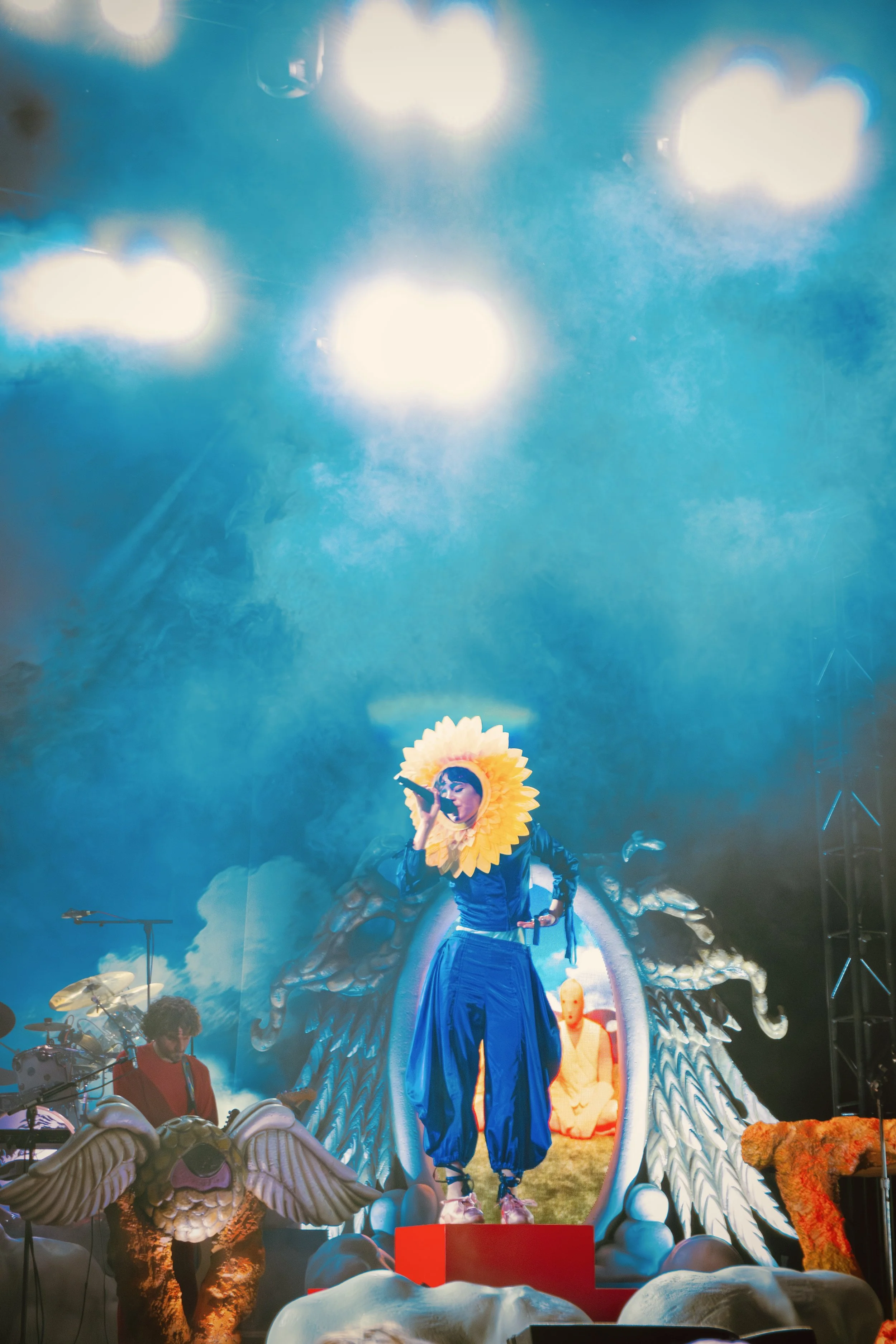 Mica Tenenbaum from Magdalena Bay on stage wearing a sunflower headpiece and a blue outfit, holding a microphone with decorative wings and clouds as part of the set design. Bright stage lighting.