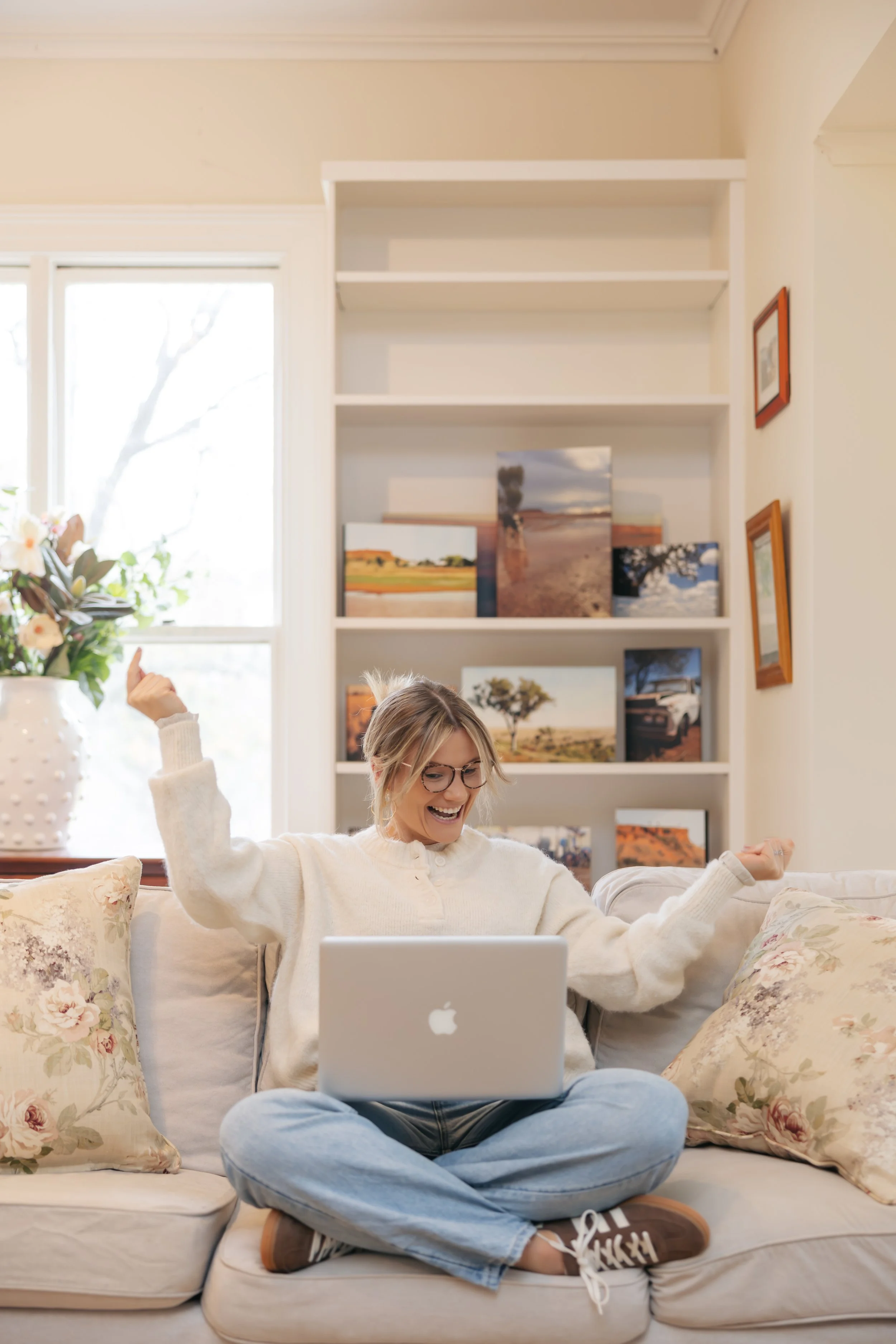 A woman sitting on a sofa with floral cushions, smiling and looking at a silver laptop on her lap, in a cozy living room with shelves and framed pictures in the background.