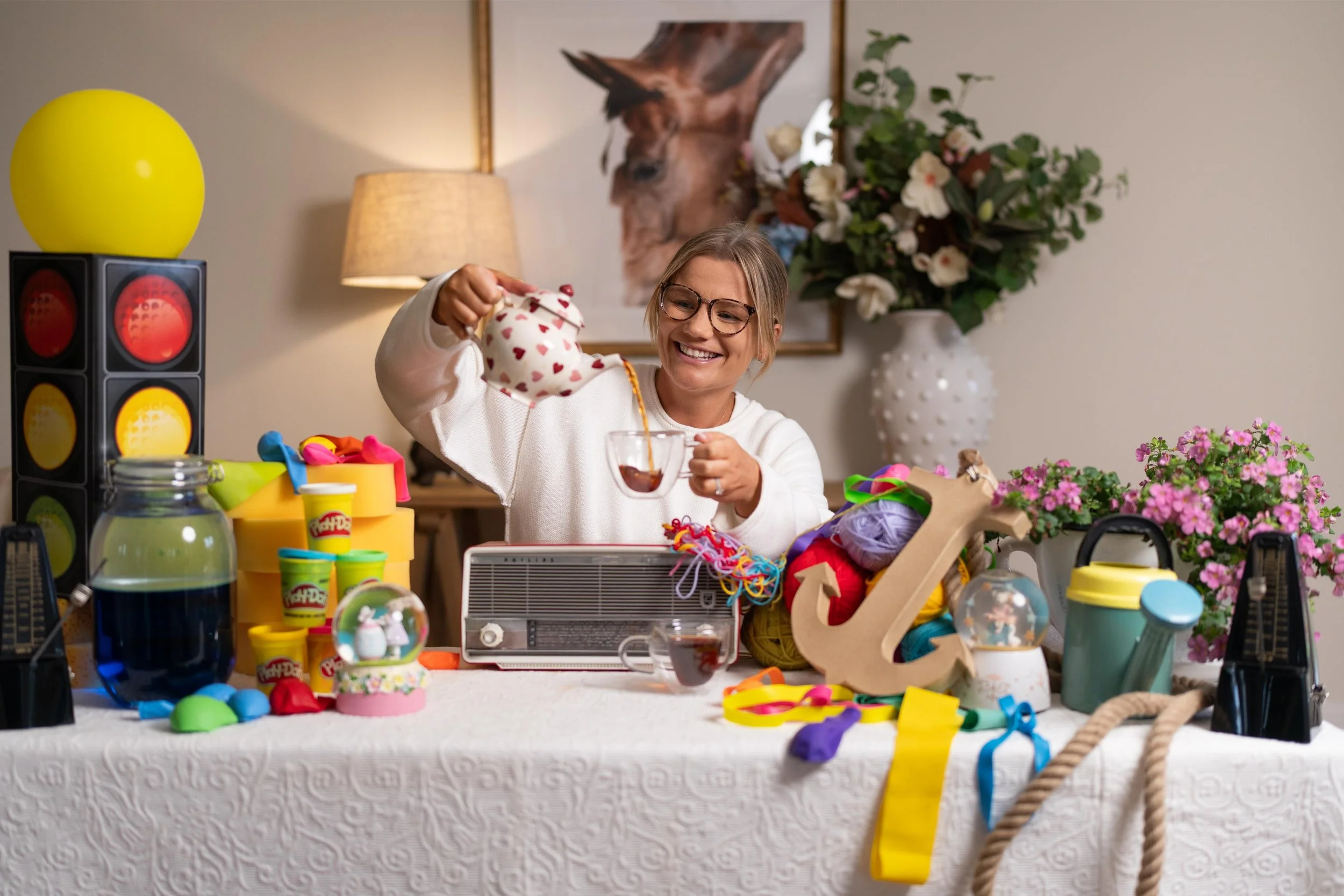 A woman with glasses smiling, holding a heart-patterned item over a glass of dark liquid, sitting at a table covered with colorful toys, craft supplies, and decorations, with a large flower arrangement and a horse portrait in the background.
