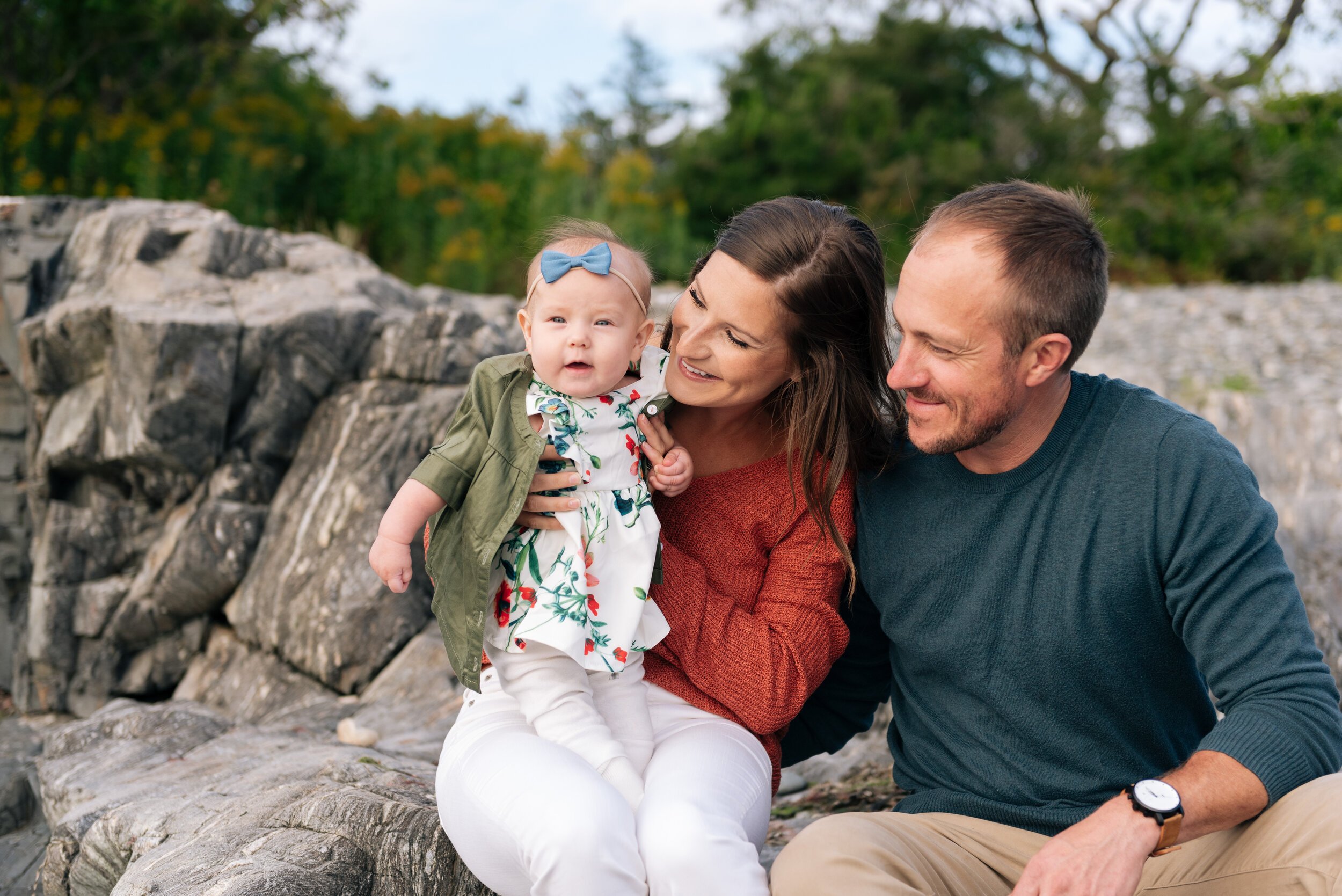 A family of three, a woman, a man, and a young girl, sitting on rocks outdoors, smiling and enjoying time together.