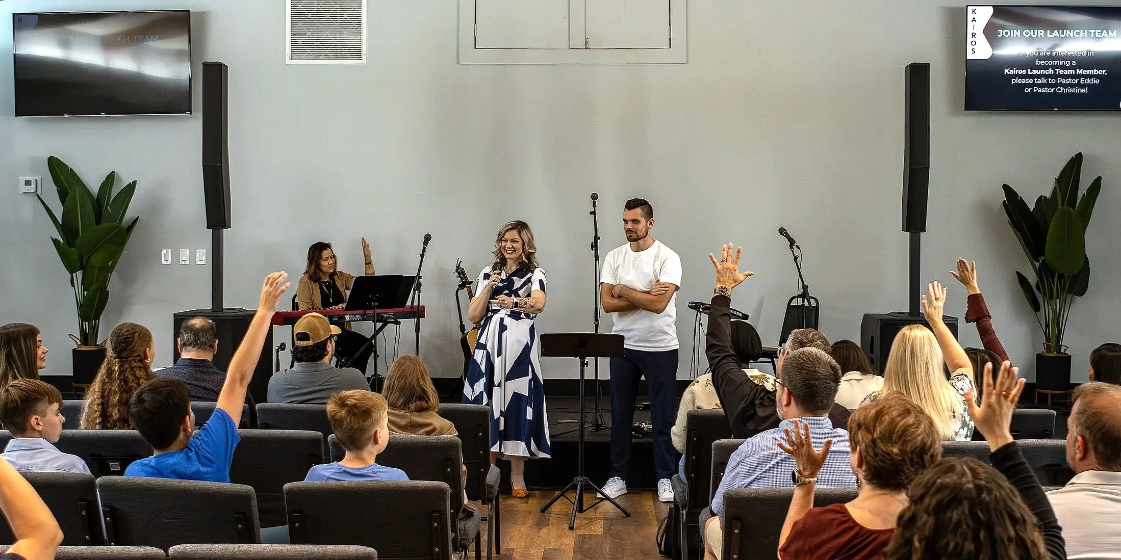 A group of people attending a church or community event, with some raising their hands. Two individuals stand at the front, one woman with a microphone and one man with crossed arms. A musician plays a keyboard on the stage, and there are large potted plants and two big speakers on either side of the stage. A screen in the background displays a message about joining the launch team.