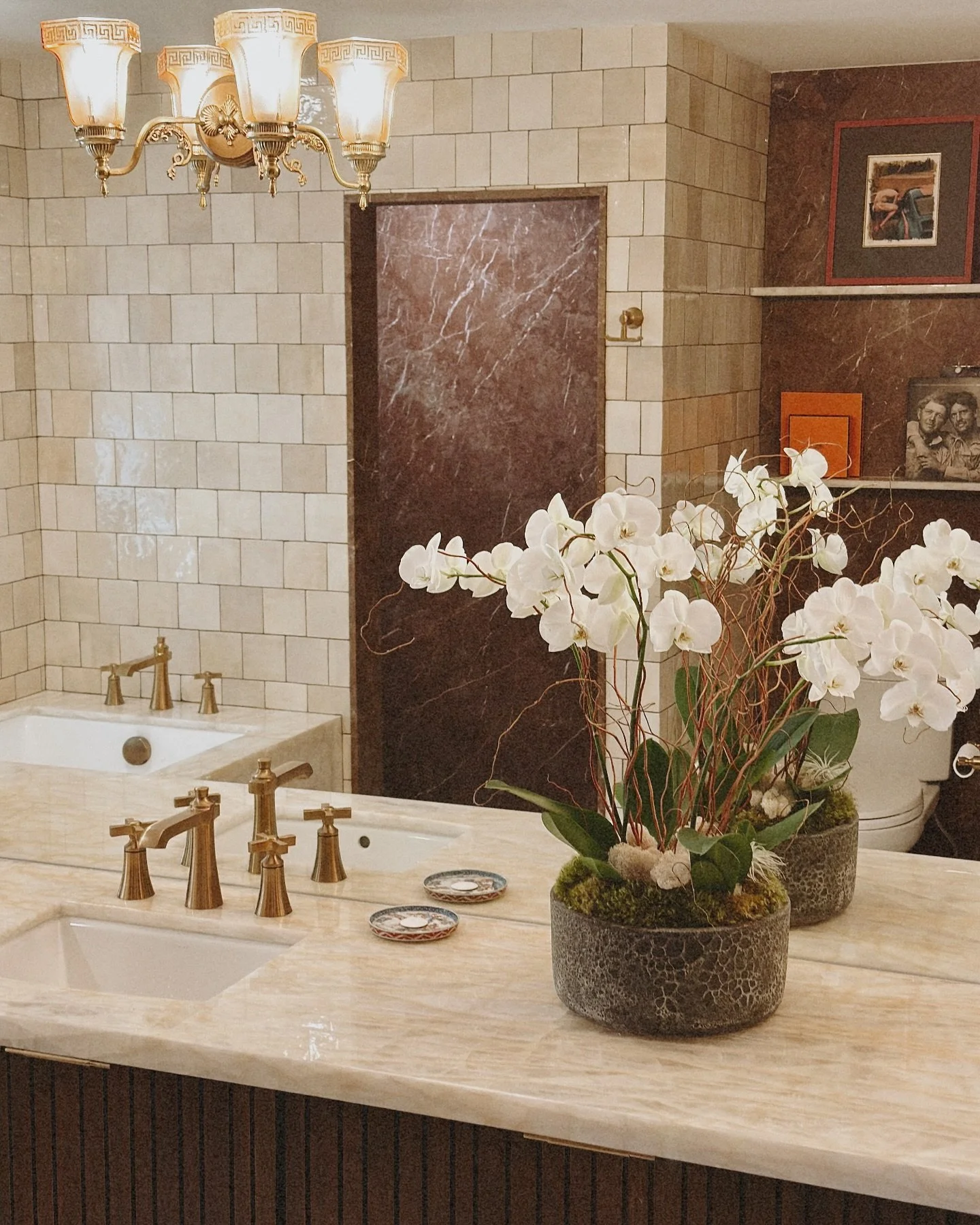 Timeless luxury meets modern function in this freshly remodeled bath retreat. From the warm brass fixtures and Grecian-inspired light fittings to the rich marble textures and serene orchid arrangement&mdash;every element was chosen to elevate the dai