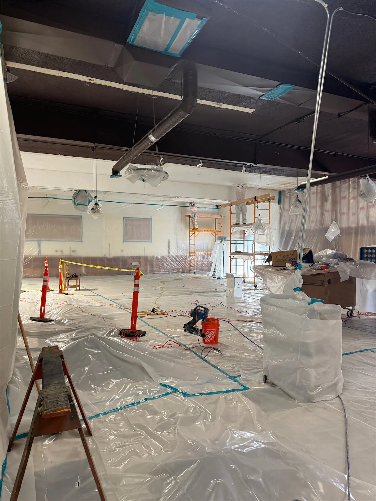 Interior of a building under renovation with plastic sheeting on the floor, scaffolding, and construction equipment.