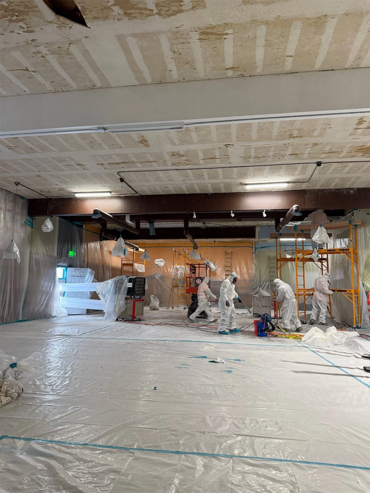 Construction workers in white protective suits working on scaffolding inside a building with plastic sheeting and temporary lighting.