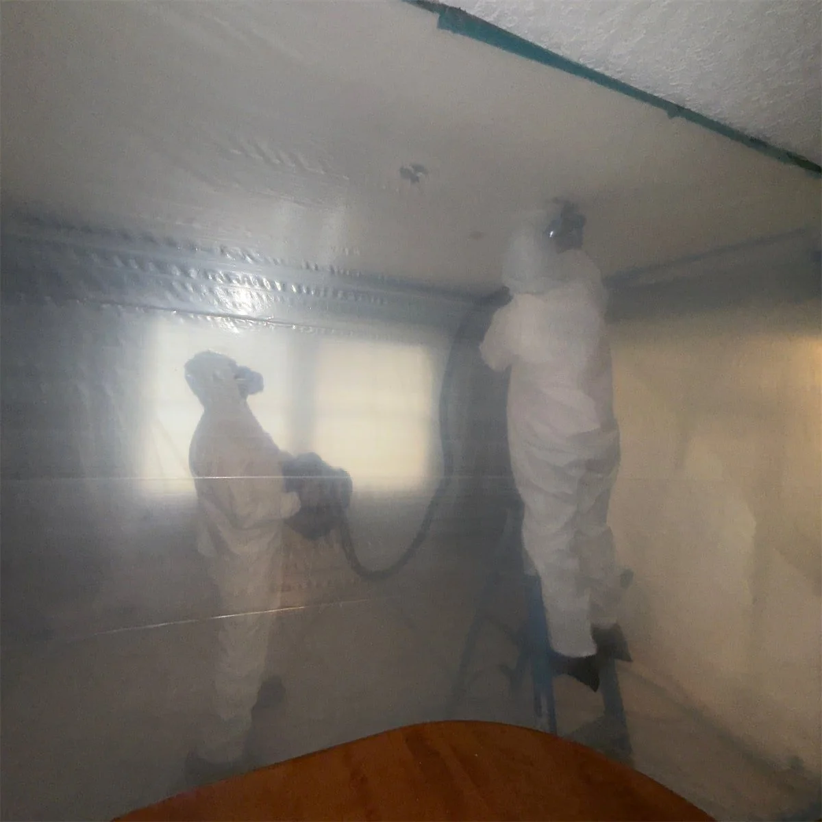 Two workers in protective suits and masks disinfect a mirror with a fogger or sprayer in a room with ornate crown molding and a window in the background.