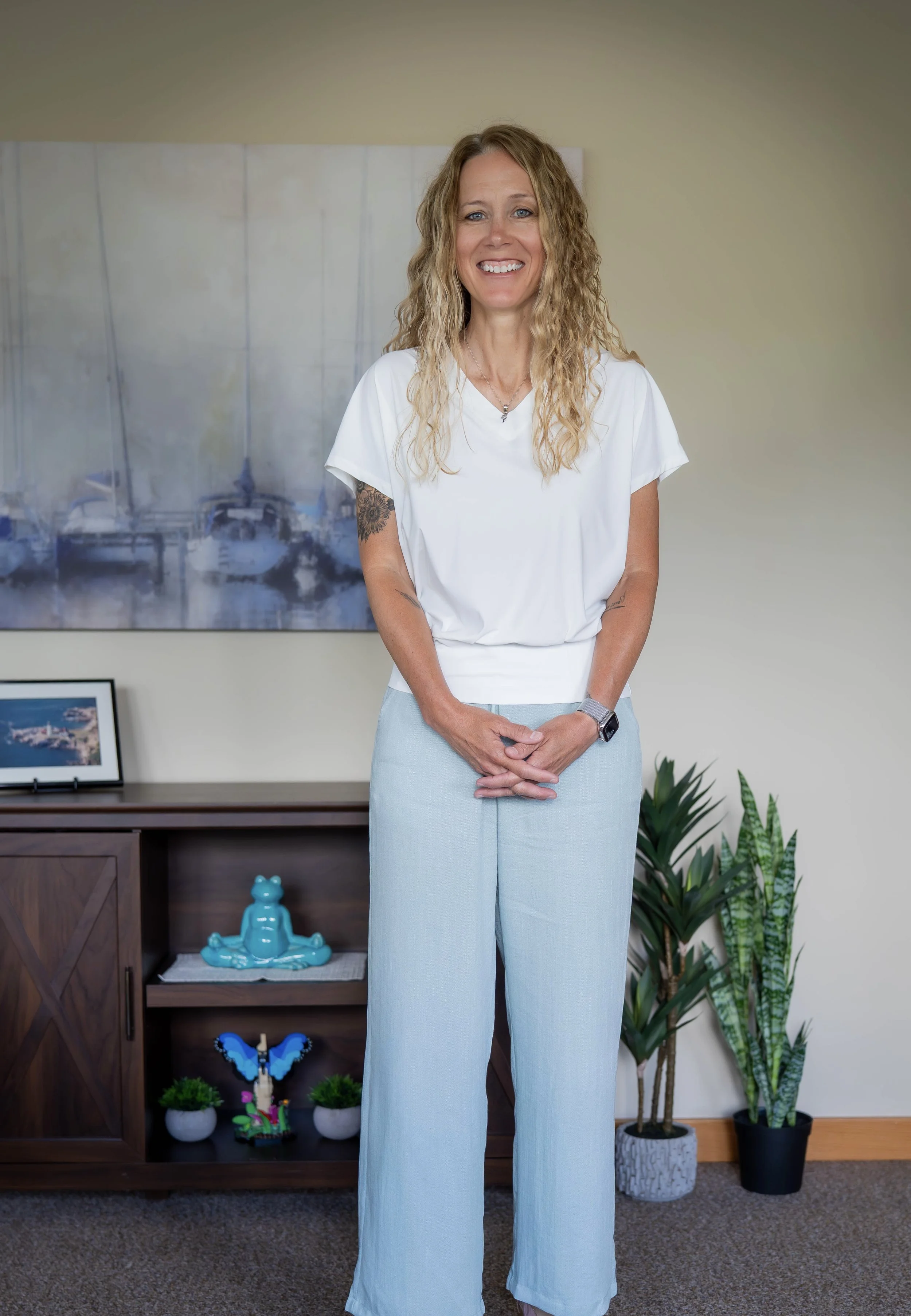 A woman with curly blonde hair smiling, wearing a white top and light blue wide-leg pants, standing in a room with plants and decorative items, including a blue butterfly sculpture and a Buddha figurine.