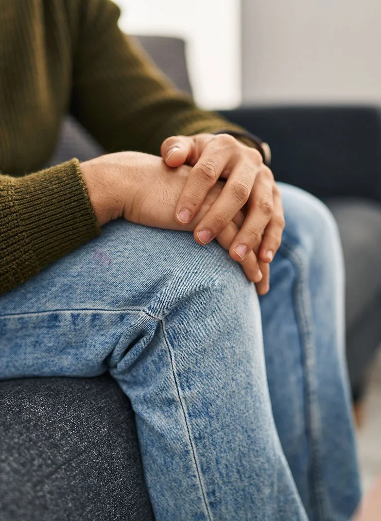 Person sitting on a dark couch wearing jeans and a brown sweater, holding their knee with both hands.