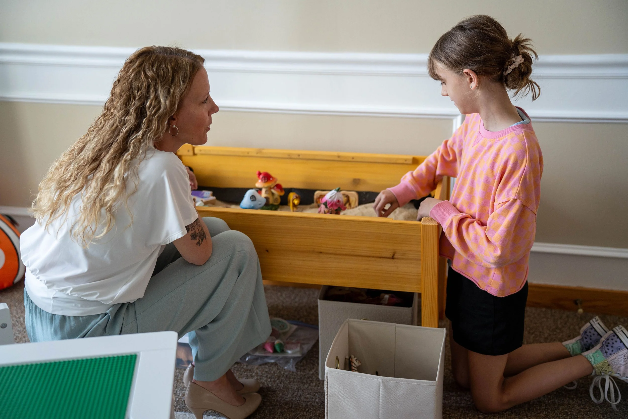 A woman sitting on the floor watching a young girl standing next to a toy bed with dolls and toys, in a room with beige walls and a carpeted floor.