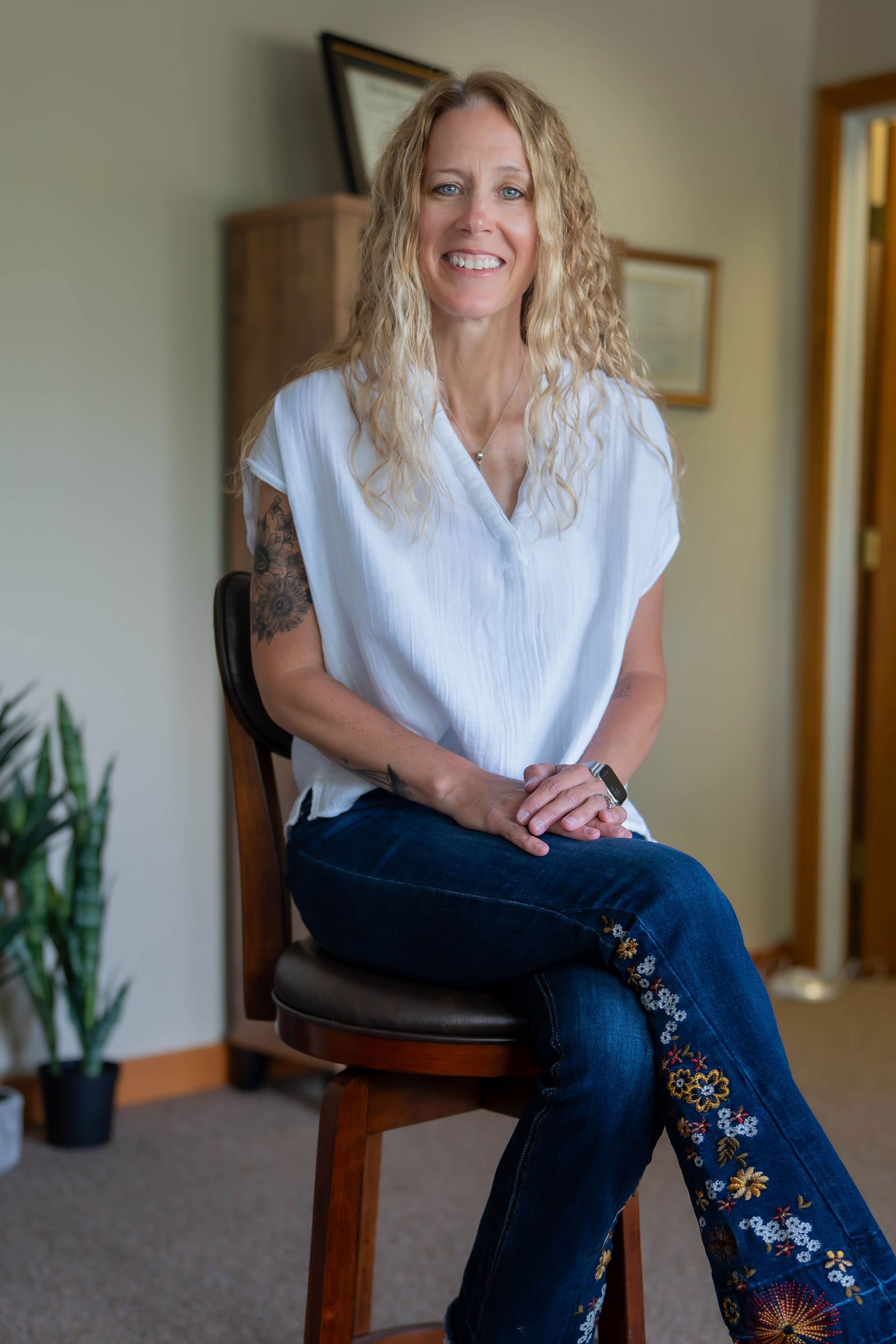 A smiling woman with curly blonde hair, wearing a white top with embroidered floral patterns on the pants, sitting on a wooden chair in a room with framed pictures and a plant in the background.