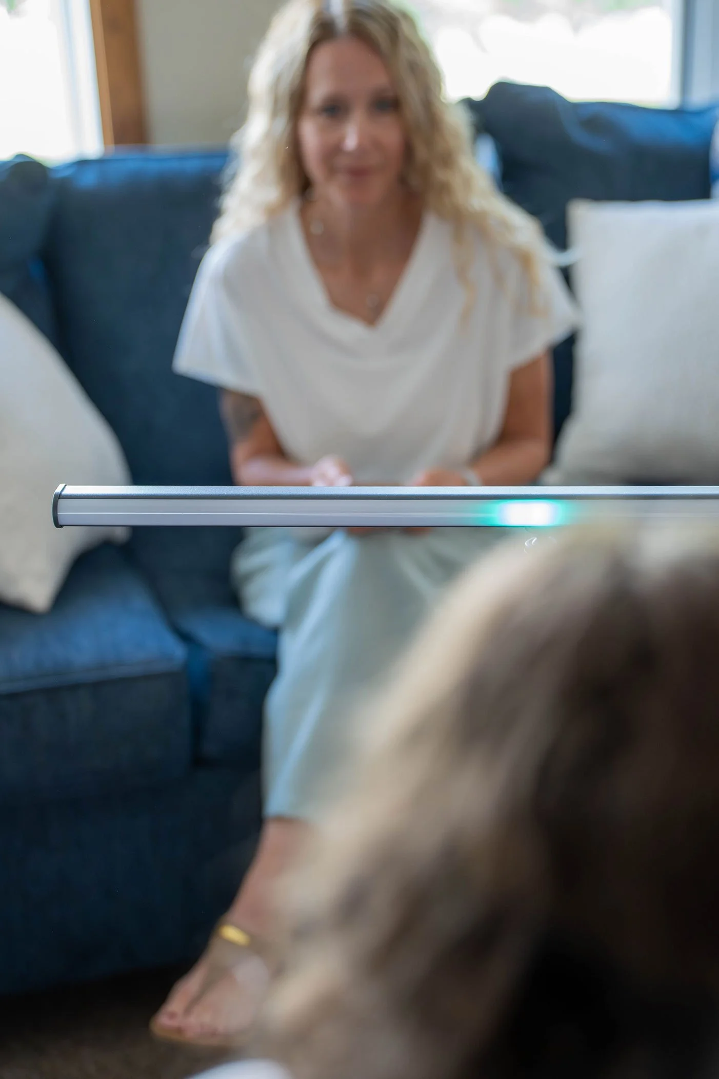 A woman with blonde curly hair sitting on a dark blue couch, blurred background, wearing a white top.