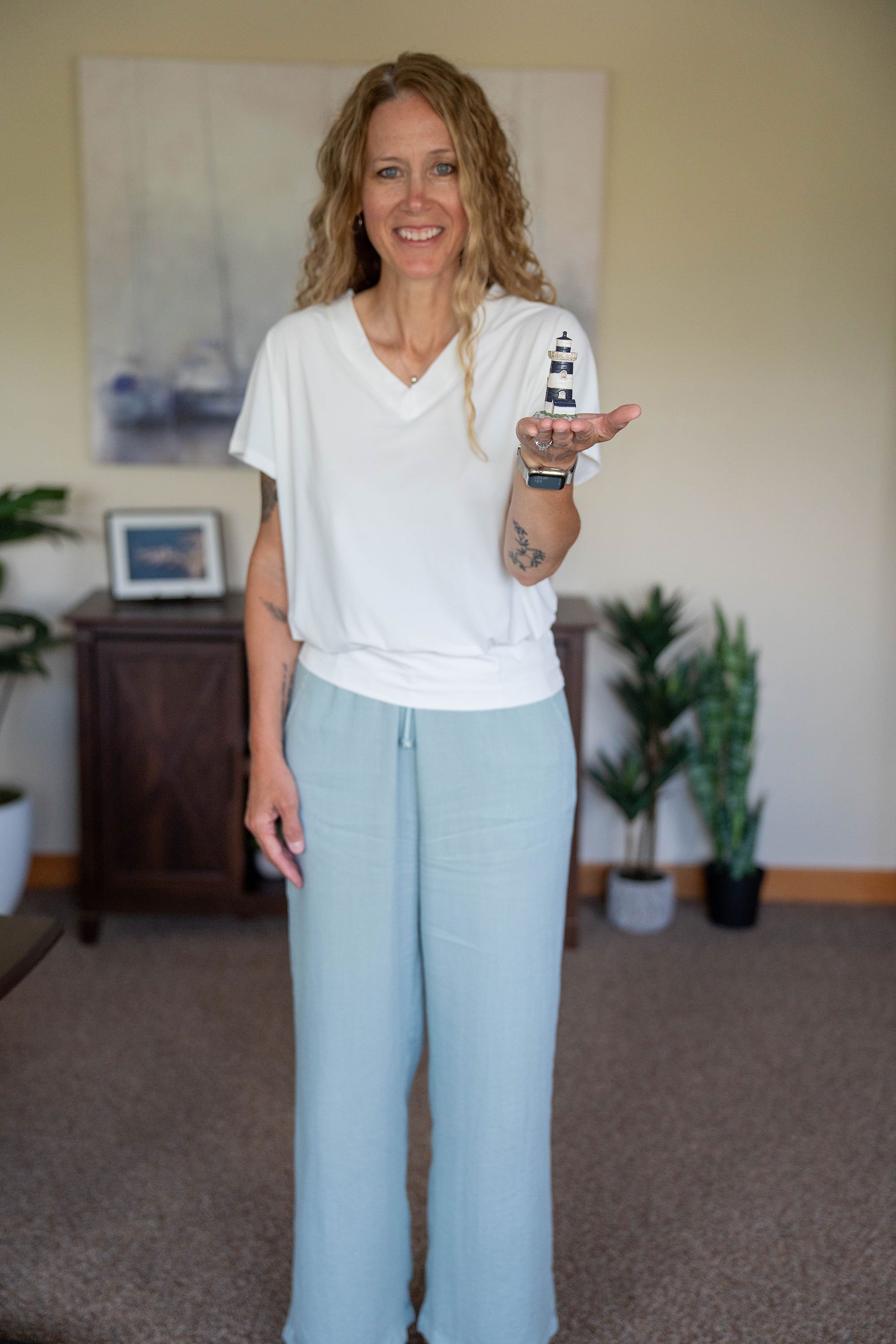 A woman with blonde, curly hair smiling while holding a miniature lighthouse model in her hand, standing in a room with potted plants and framed pictures.