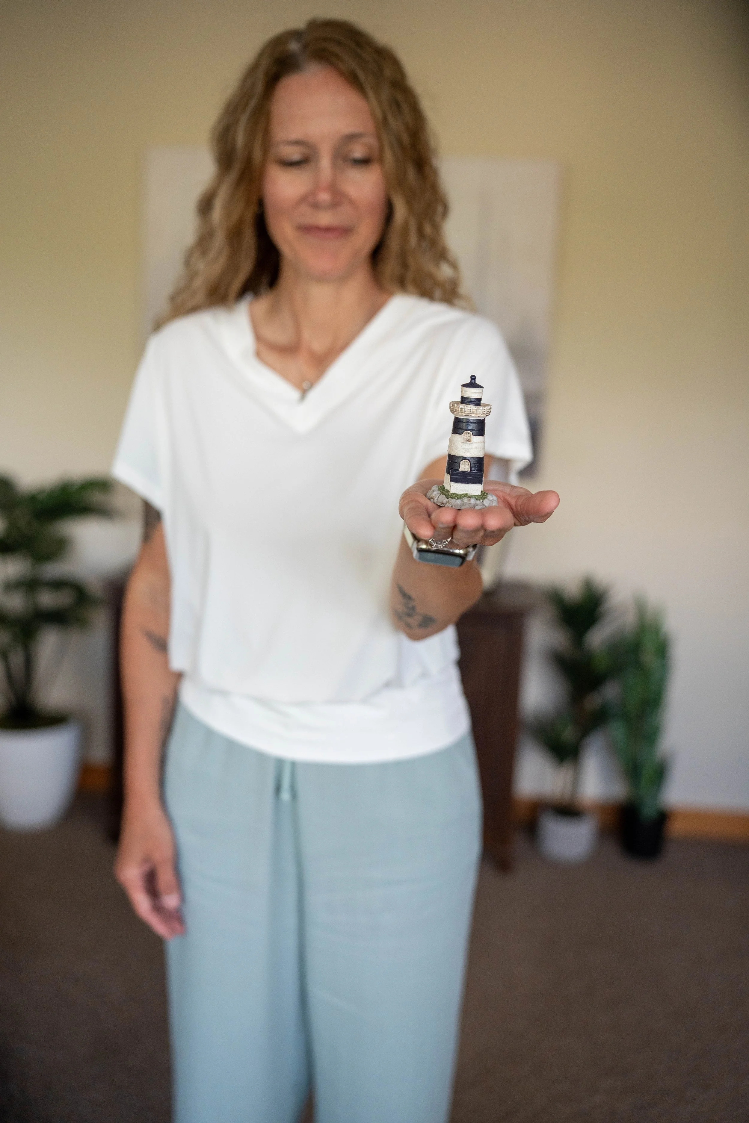 A woman with vertical wavy hair in a white T-shirt and light blue pants holding a miniature lighthouse figurine in her right hand, standing in a room with several potted plants in the background.