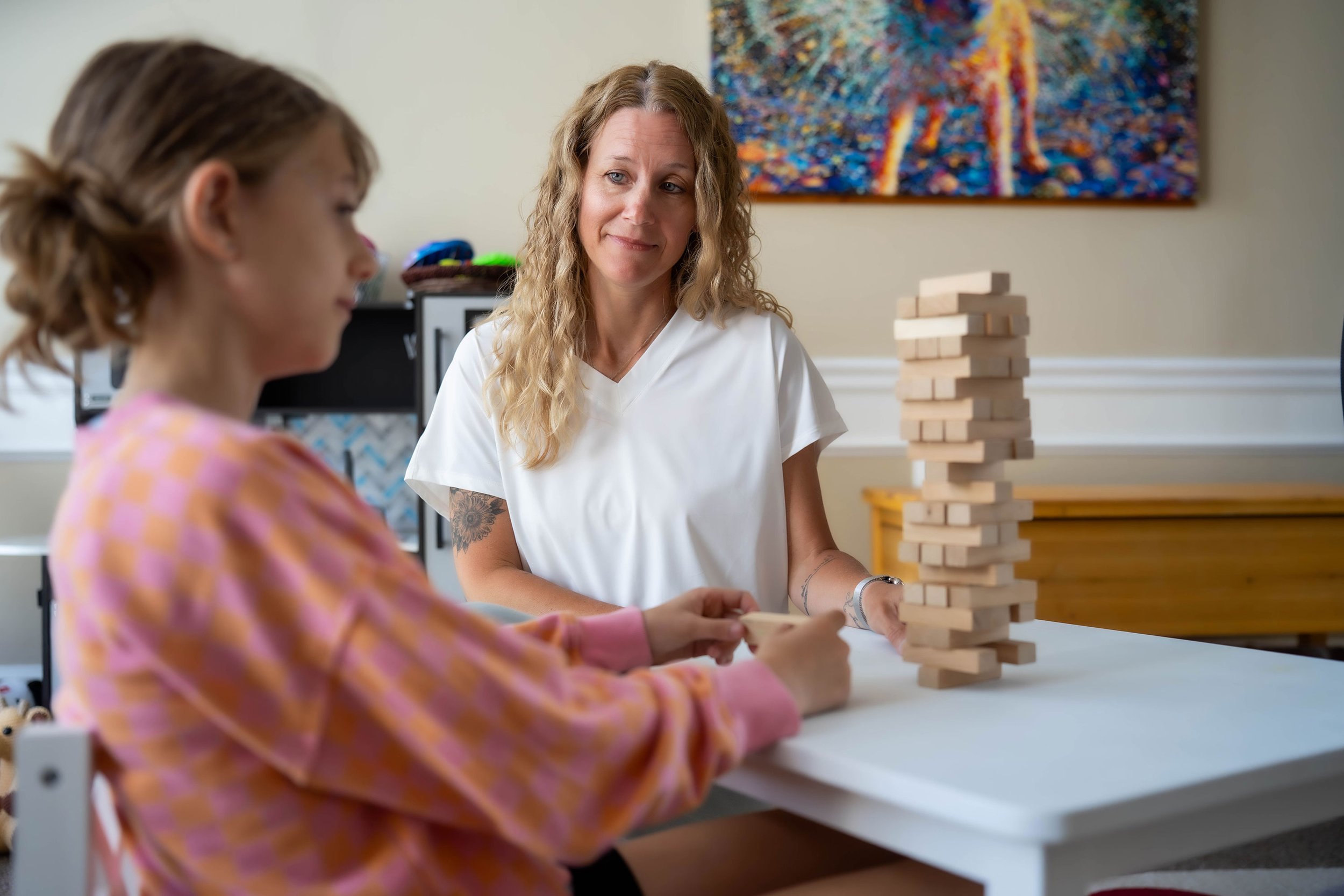 A woman and a young girl are playing a giant Jenga game at a table in a room with colorful artwork on the wall. The woman is watching the girl as she carefully removes a block from the Jenga tower.