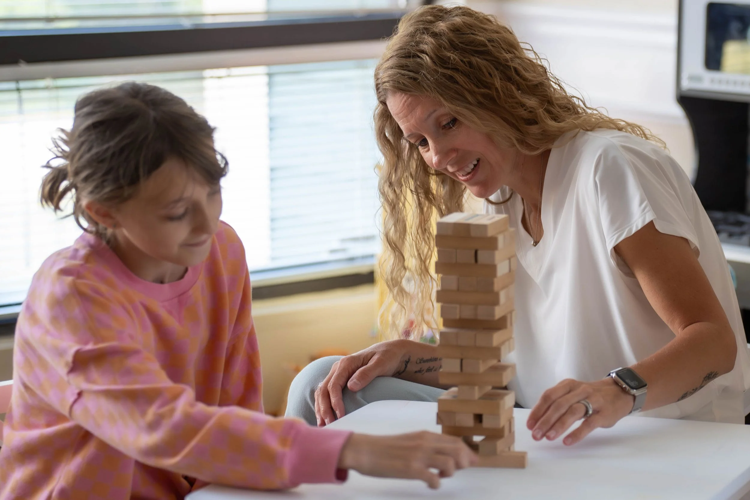 A woman and a young girl play a game of Jenga, balancing wooden blocks on a table near a window with blinds.
