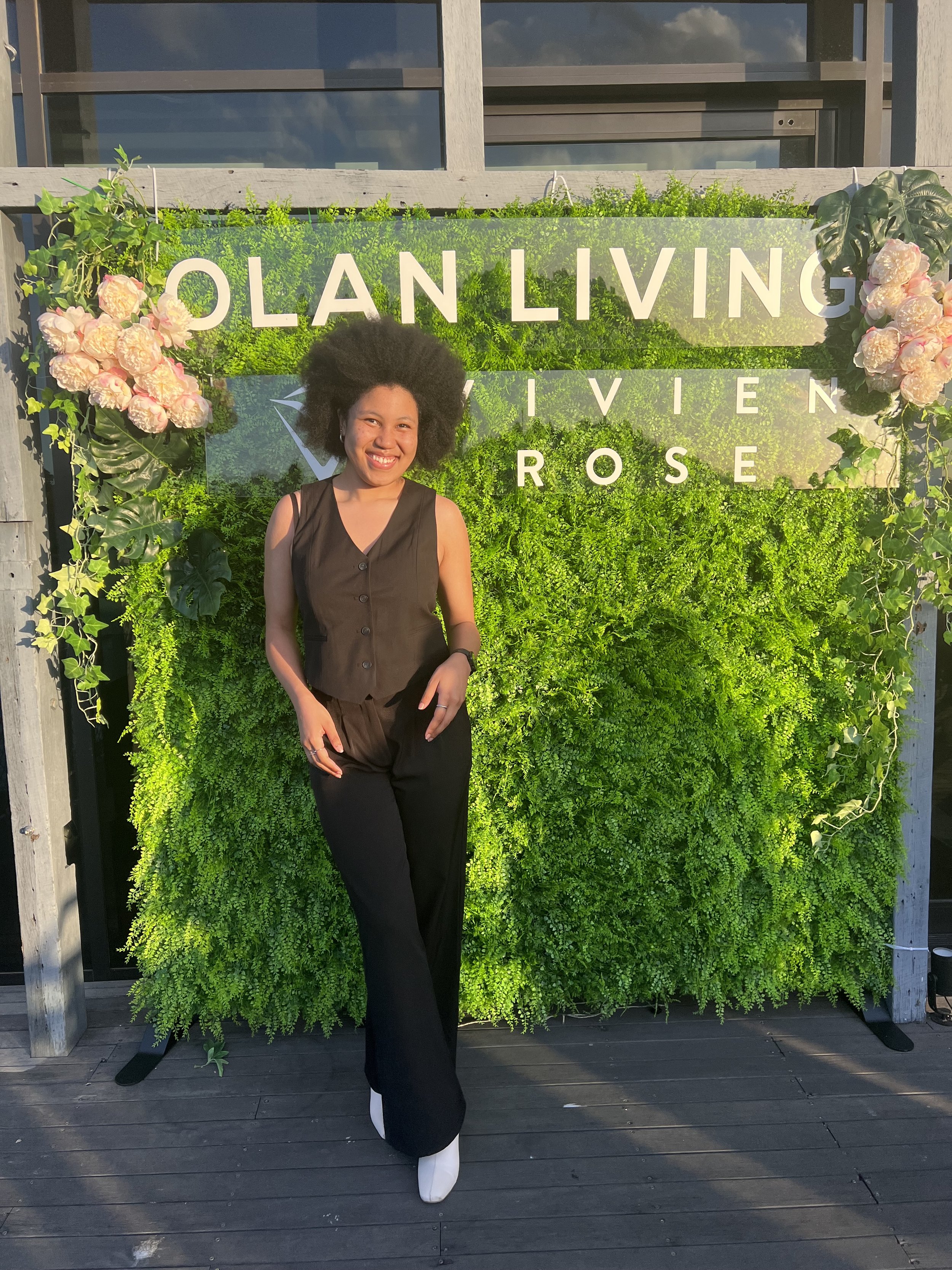 Young woman with curly hair smiling, standing in front of a green living wall with pink flowers and a sign that reads 'Olan Living, Vivian Rose', on a wooden deck.