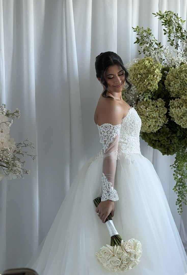 A bride in a white wedding gown holding a bouquet of white roses, standing in front of a white curtain and surrounded by green and white floral arrangements.