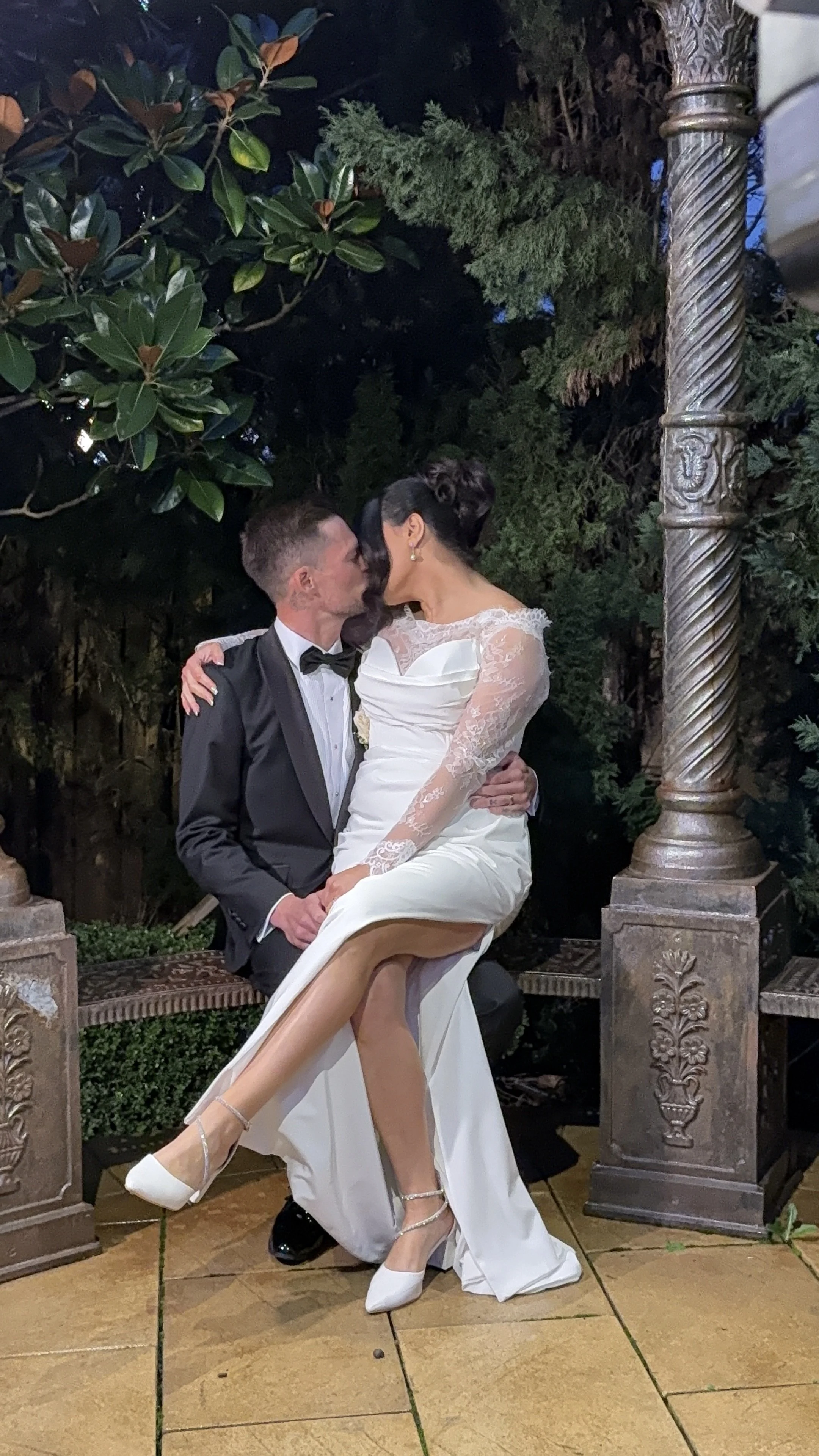A newlywed couple kissing on a garden bench at night, the bride in a white wedding gown with lace sleeves and high heels, the groom in a black tuxedo with a bow tie, surrounded by lush greenery and decorative stone columns.