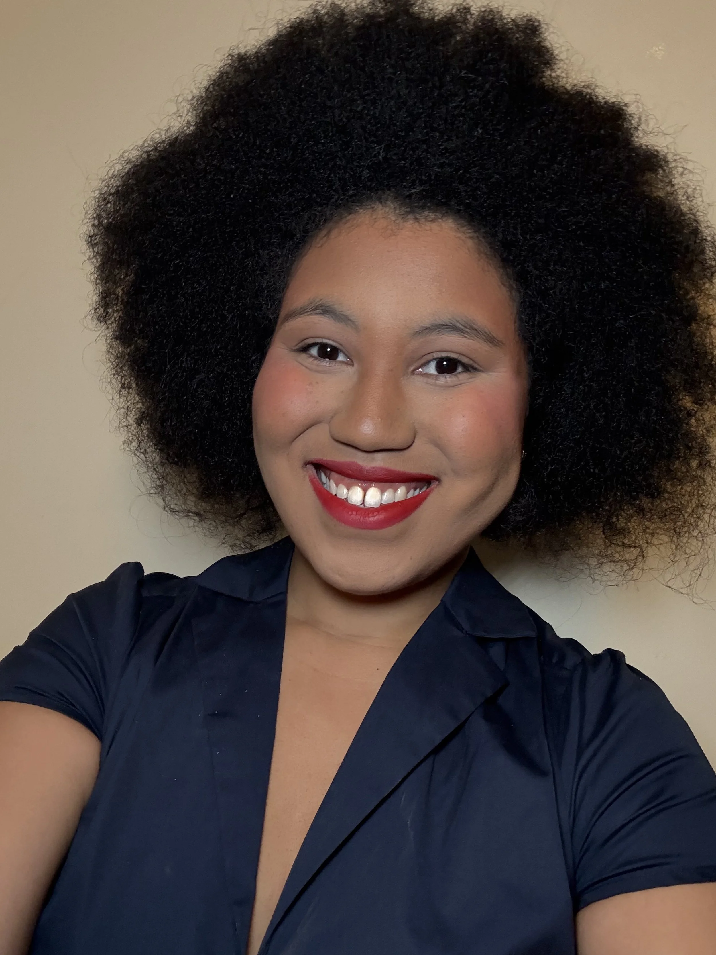 A smiling woman with natural curly hair, wearing red lipstick and a navy blue top, taking a selfie against a plain beige wall.