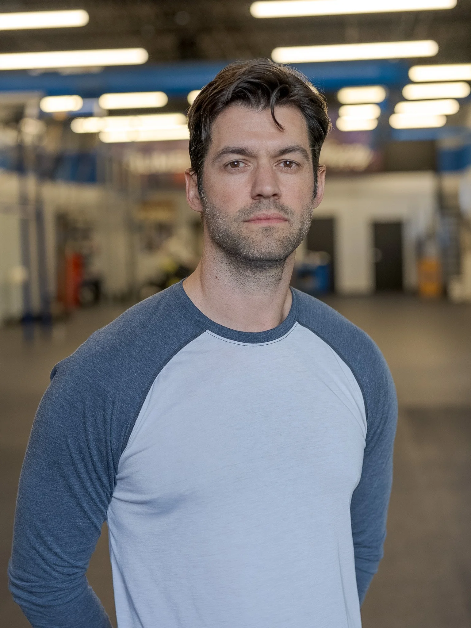 A man with dark hair, beard, wearing a gray and white baseball tee, standing in an indoor gym or warehouse with bright overhead lighting.