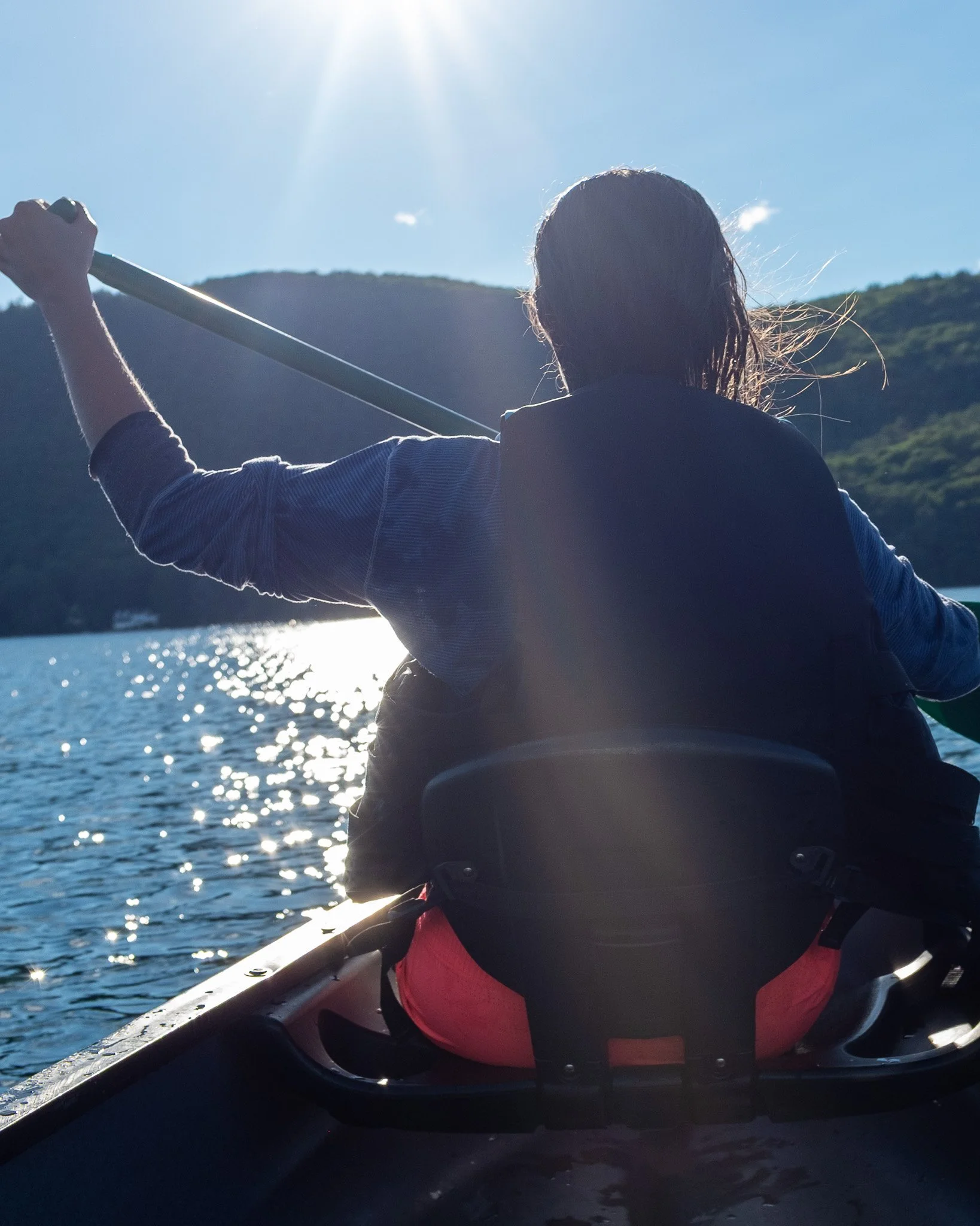 Person kayaking on a sunny water body, back view, with mountains in the background.