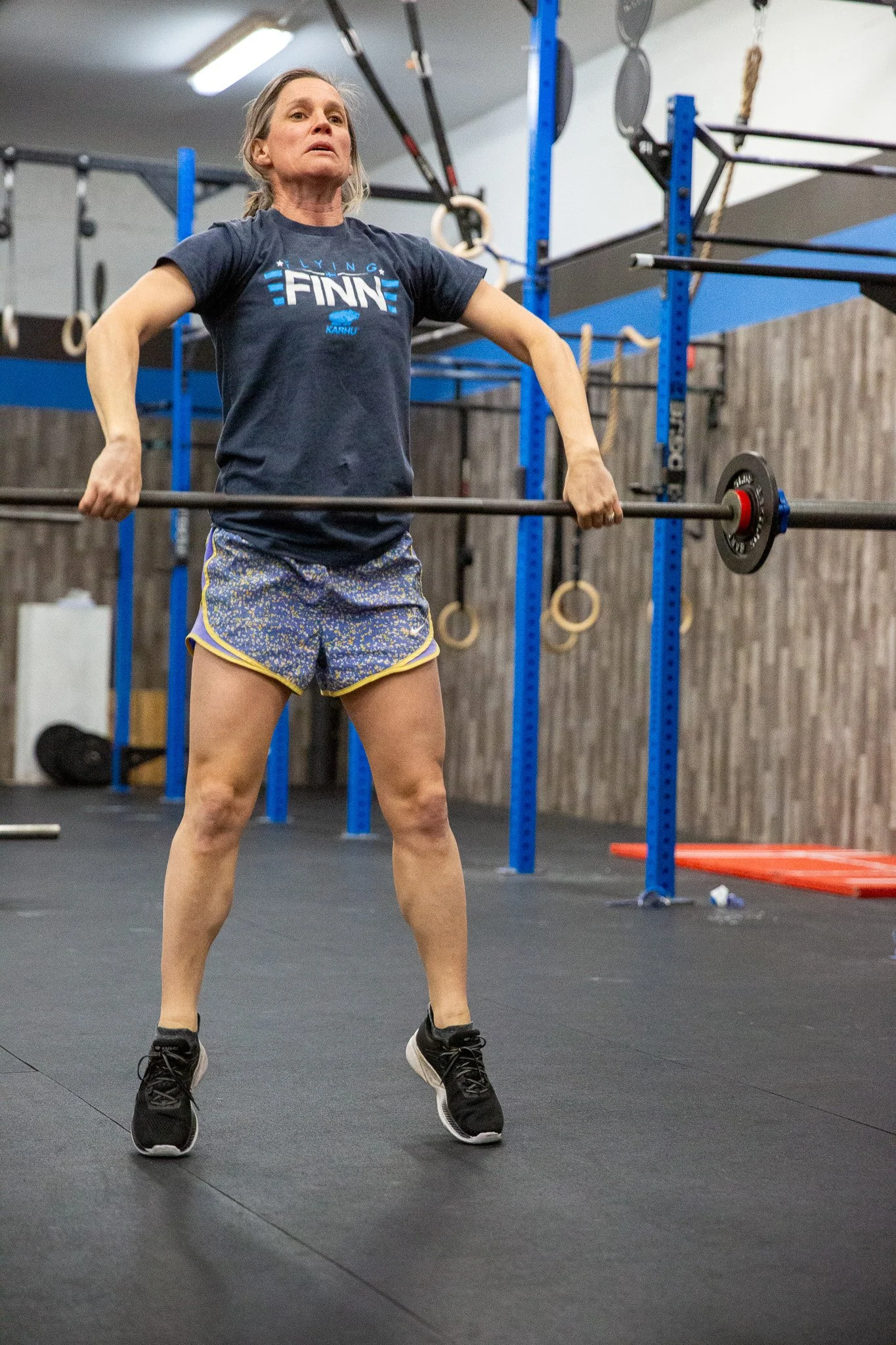 A woman lifting a barbell in a gym with gymnastic rings and blue wall-mounted equipment in the background.