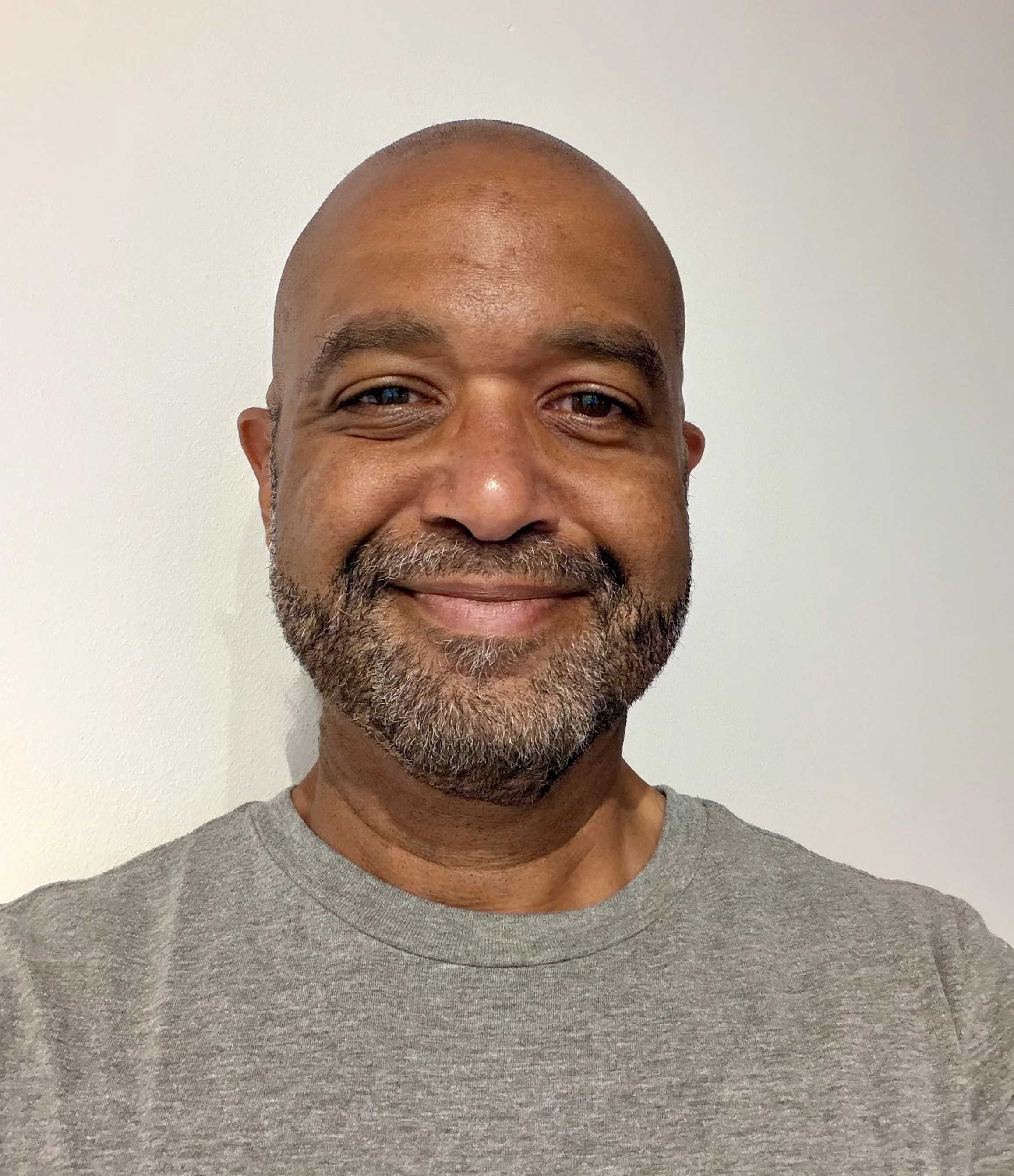 Close-up of a smiling man with a bald head and beard, wearing a gray t-shirt, standing in front of a plain white wall.