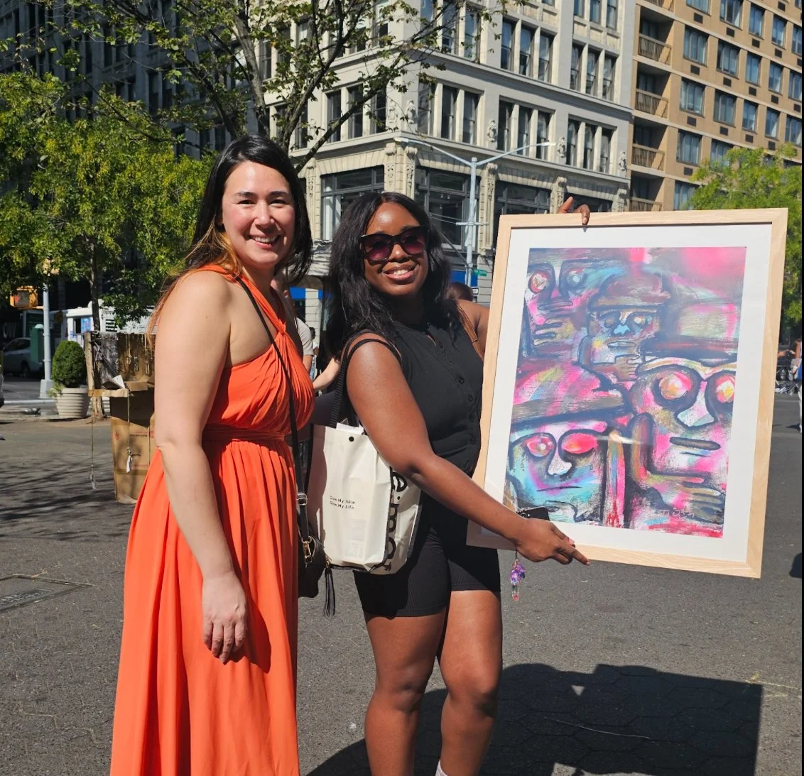 Two women smiling, standing outdoors in front of tall buildings, with one holding an abstract colorful painting of faces and figures.