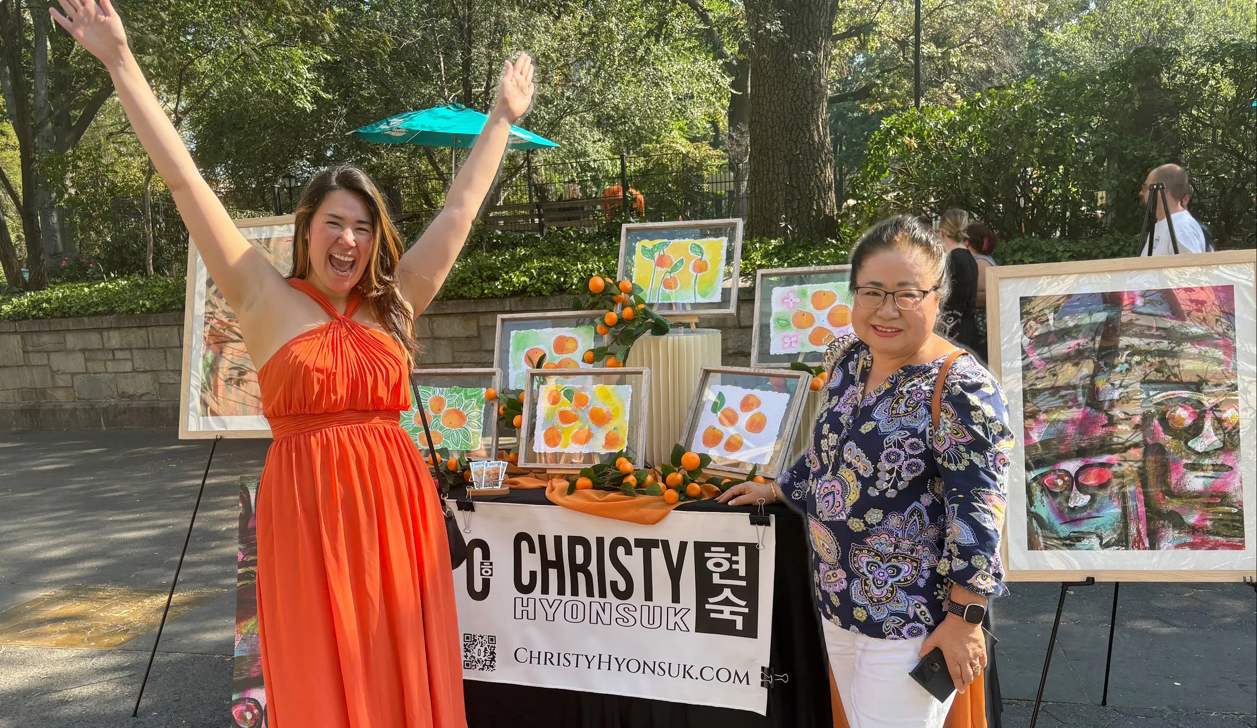 Two women at an outdoor art booth with colorful paintings and decorations, smiling and posing for the camera, with trees and other people in the background.