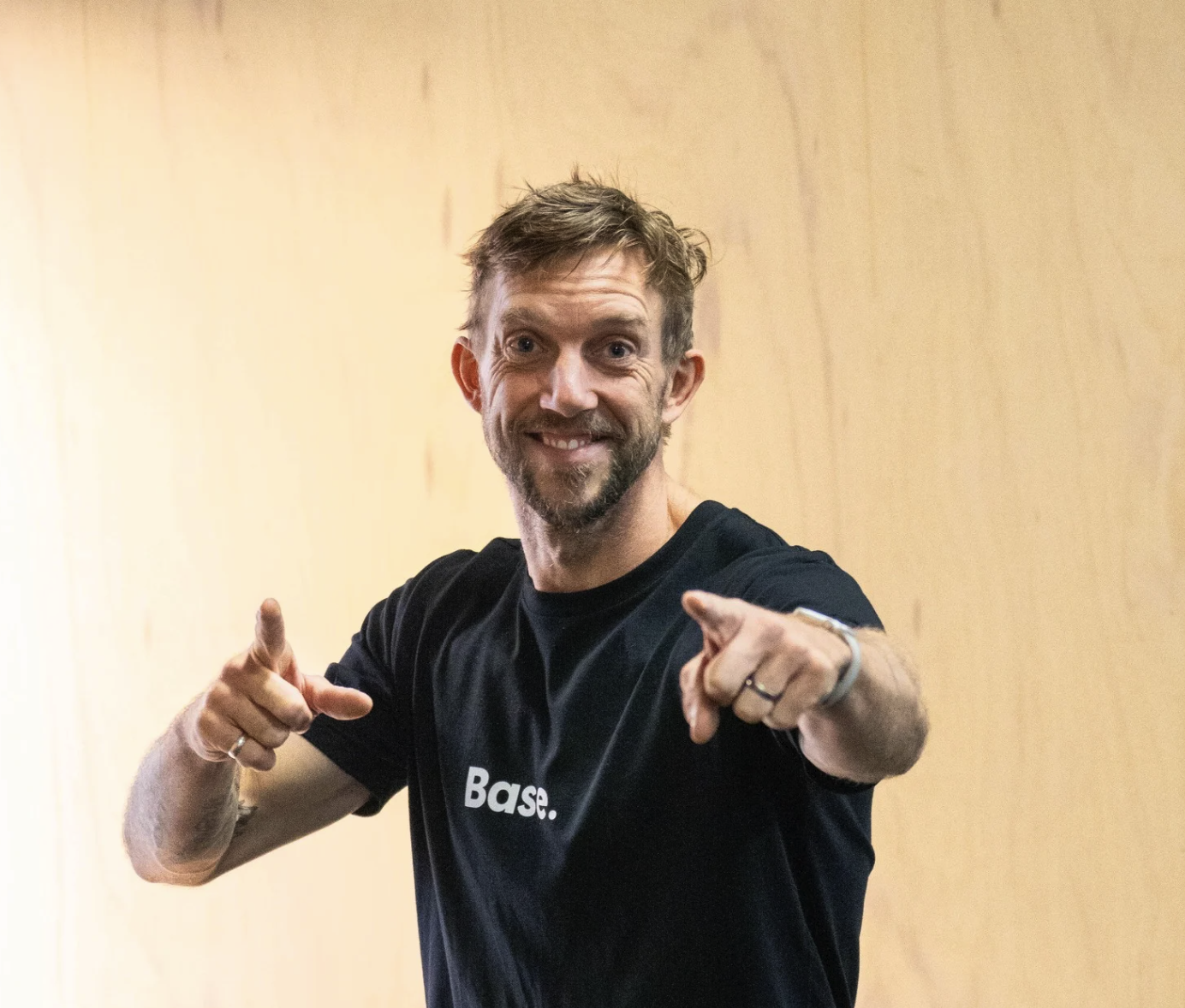 A smiling man with a beard and short brown hair wearing a black t-shirt that says "Base." pointing towards the camera in front of a plain beige wall.