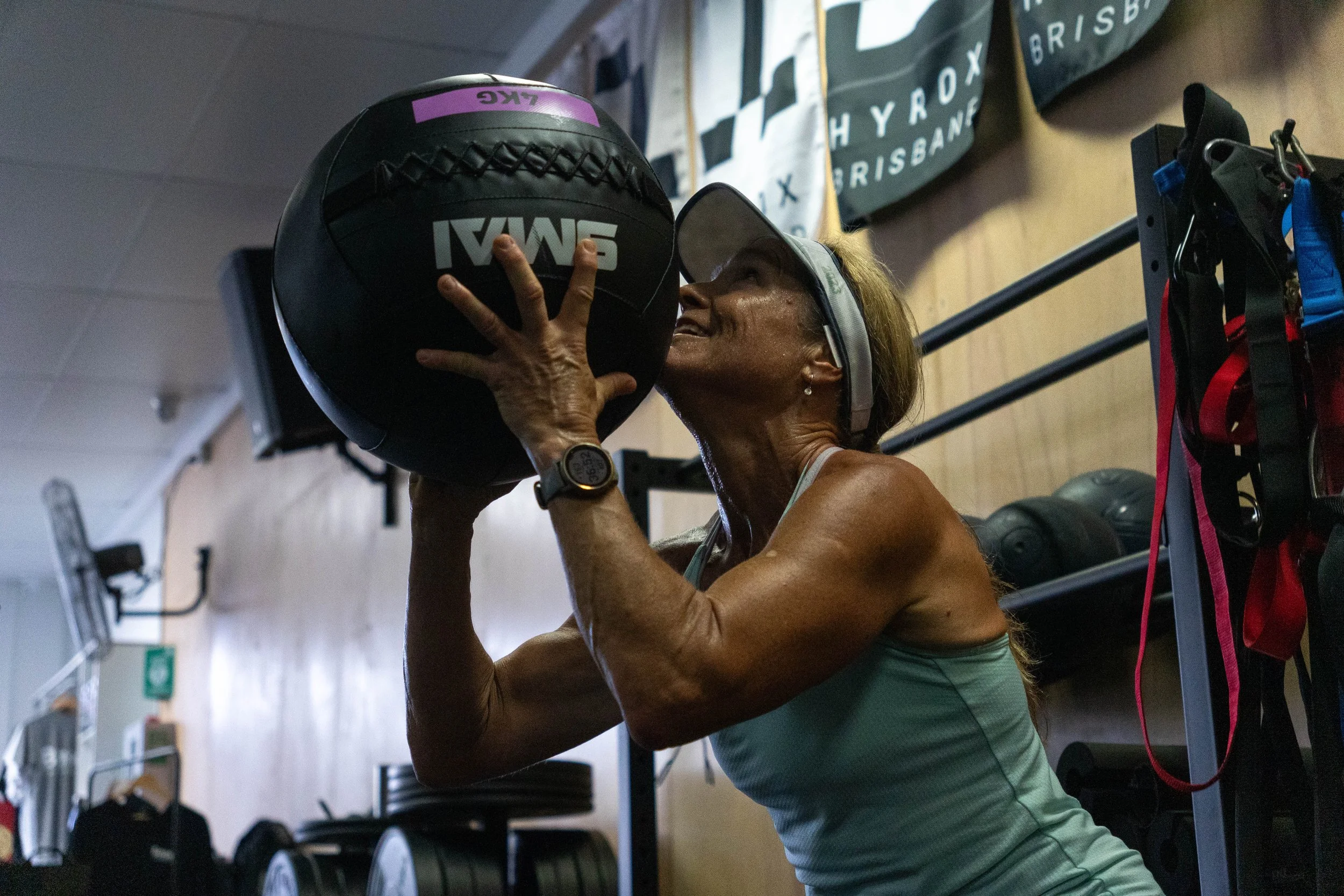 A woman wearing a visor, a fitness watch, a tank top, and earrings is lifting a black and purple medicine ball in a gym or fitness studio.