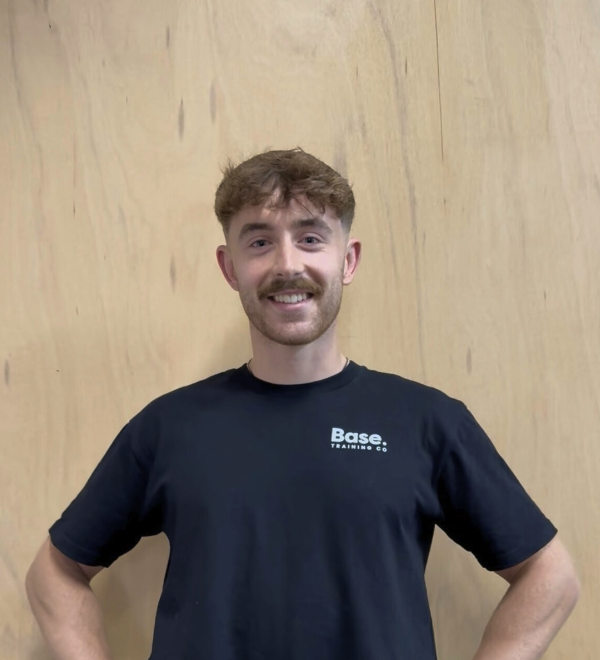 A young man with brown hair, a beard, and a big smile, wearing a black t-shirt with white text, standing in front of a wooden wall.