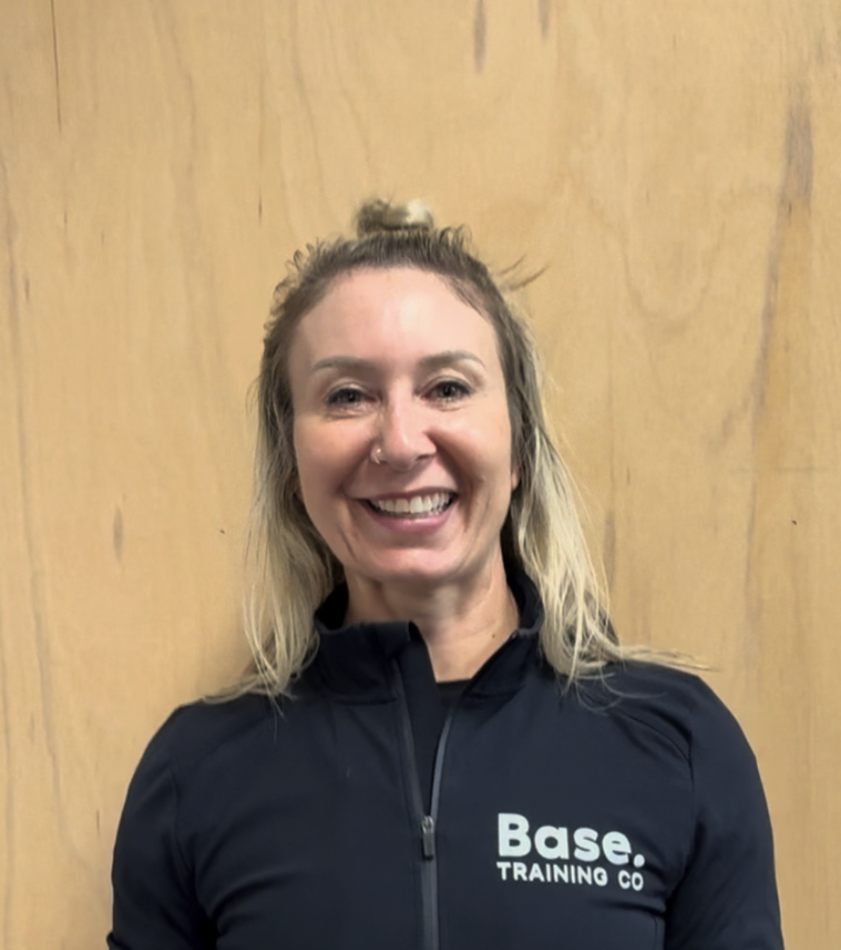 A woman smiling, wearing a black zipped sports jacket with 'Base. TRAINING CO' written on it, standing against a wooden background.