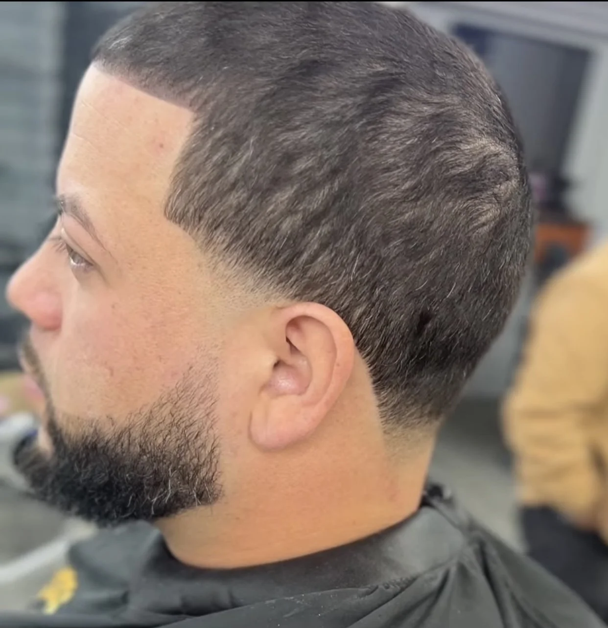 Close-up of a man's side profile with a neat, low fade haircut and well-groomed beard in a barbershop setting.