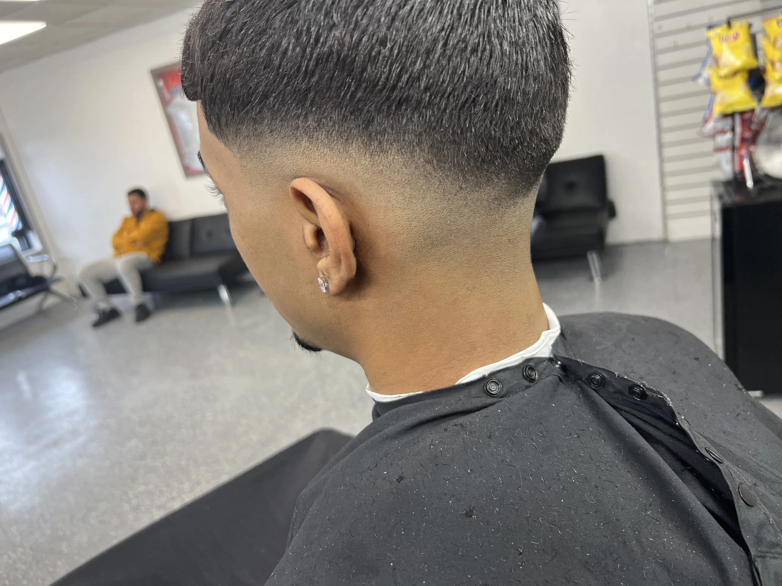 Man with a fresh fade haircut facing away in a barbershop, another person seated in the background.