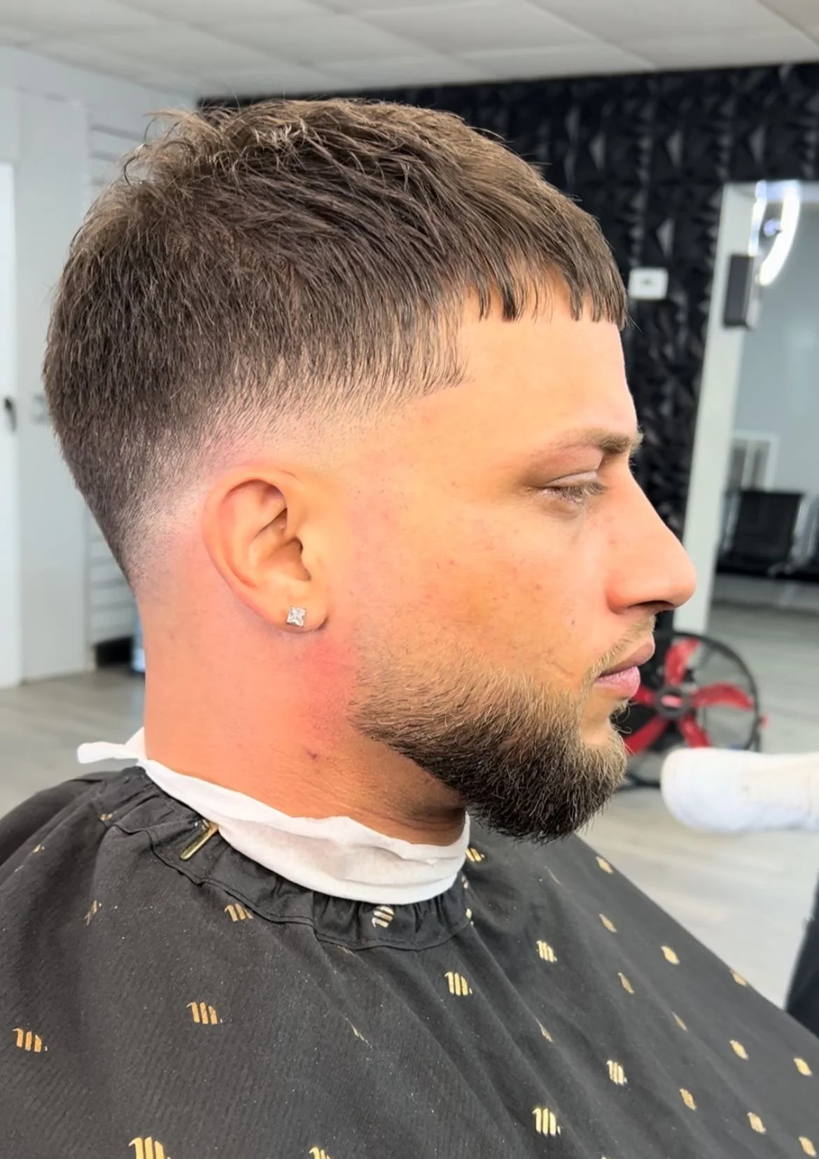 Side profile of a man with a fresh haircut, featuring a short fade and neatly trimmed beard, wearing a black barber cape and an earring.