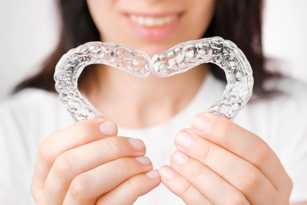 Person holding a clear dental retainer shaped like a heart with a smiling face in the background.