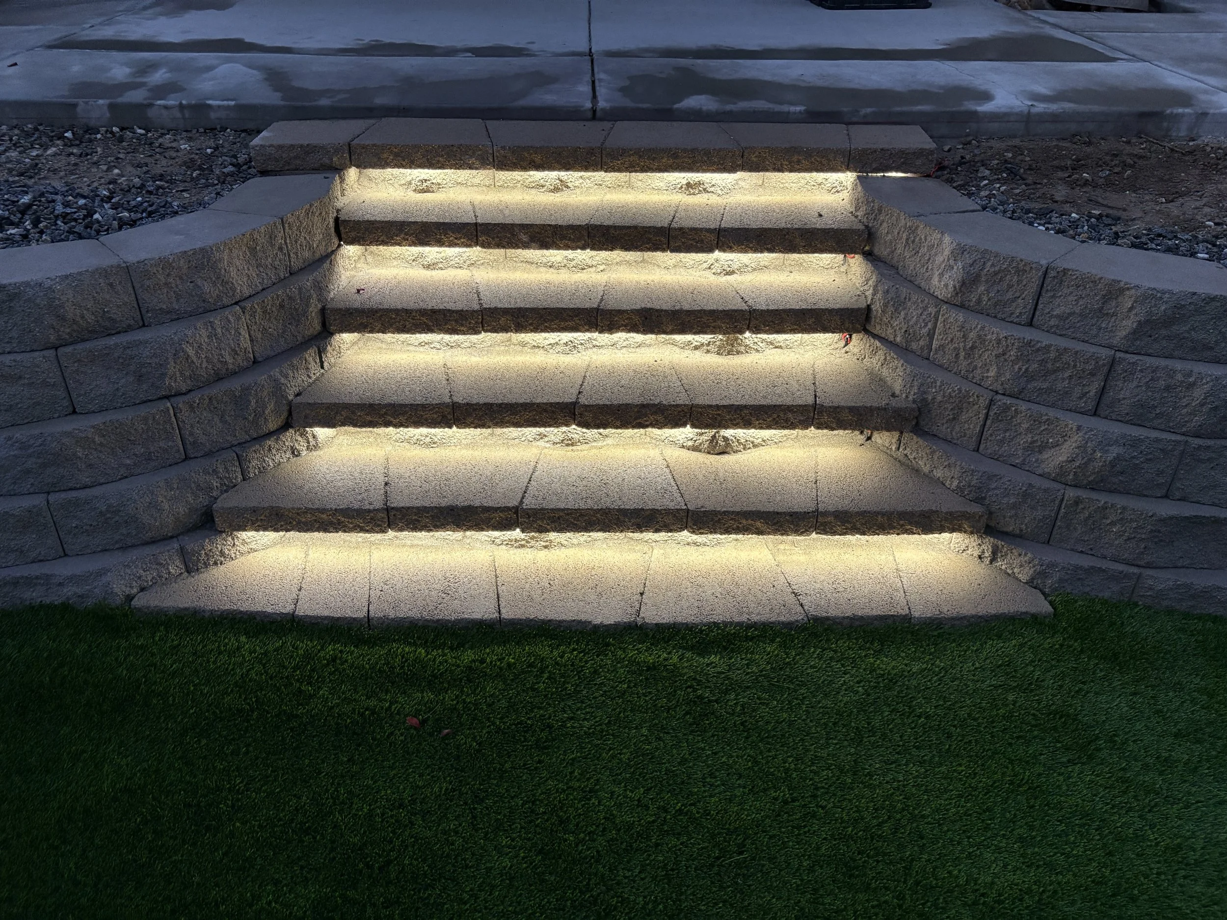 Stone staircase with built-in lighting, bordered by curved stone walls, leading to a higher level, with green grass at the bottom. Residential hardscape and landscape remodel in Canyon Lake, CA, combining stone, turf, and lighting.