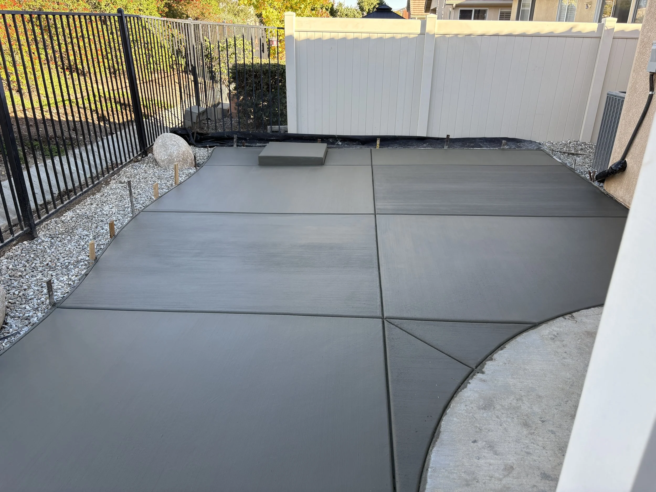 Freshly poured concrete patio with sealing joints, bordered by a curved edge, with a black metal fence on the left and a white fence in the background, and neighboring house wall on the right.