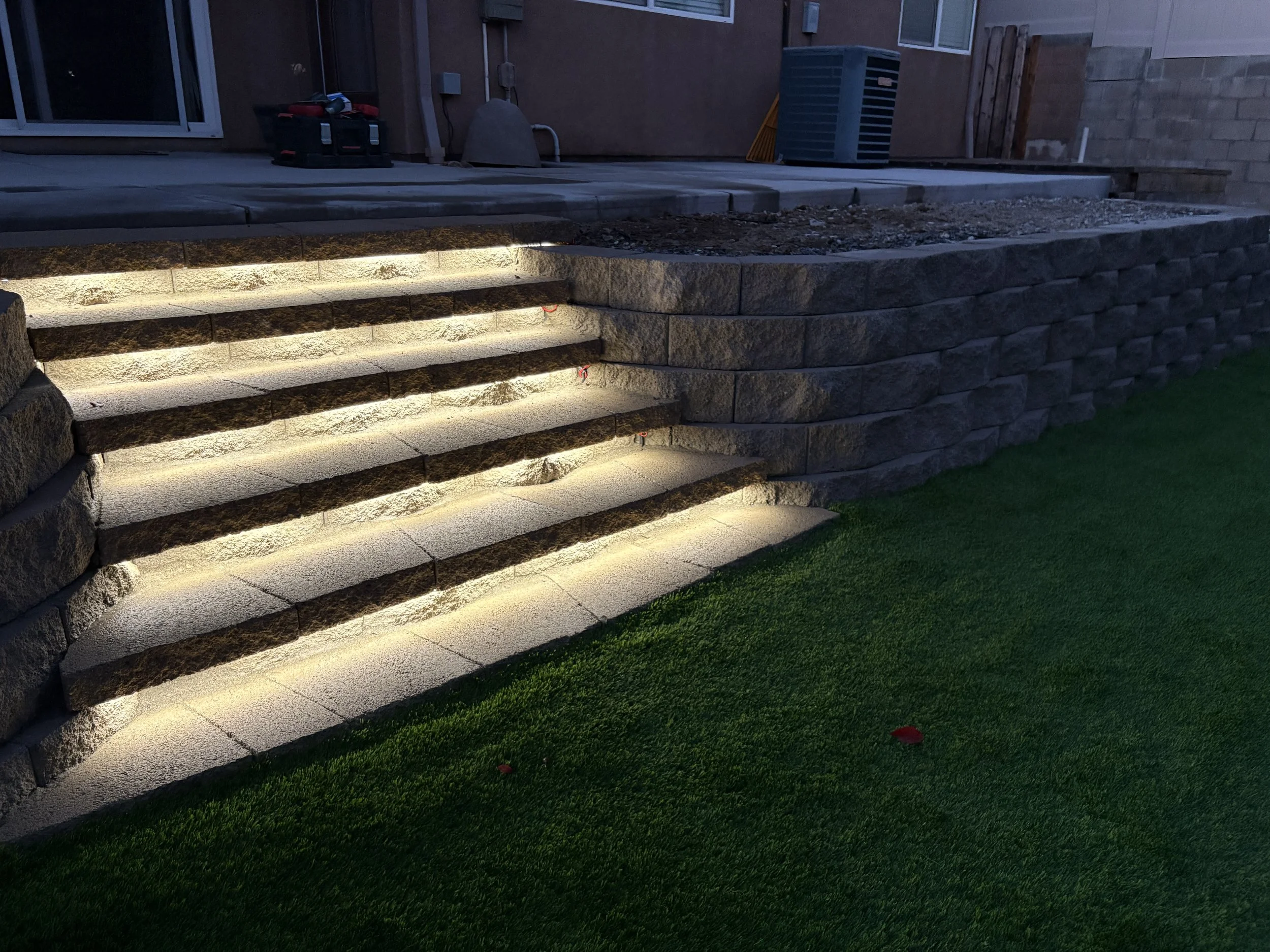 Exterior stairs leading up to a patio, illuminated by built-in lighting along each step, with a stone retaining wall and artificial grass lawn in the foreground. Canyon Lake yard upgrade featuring illuminated stone steps and premium artificial turf.
