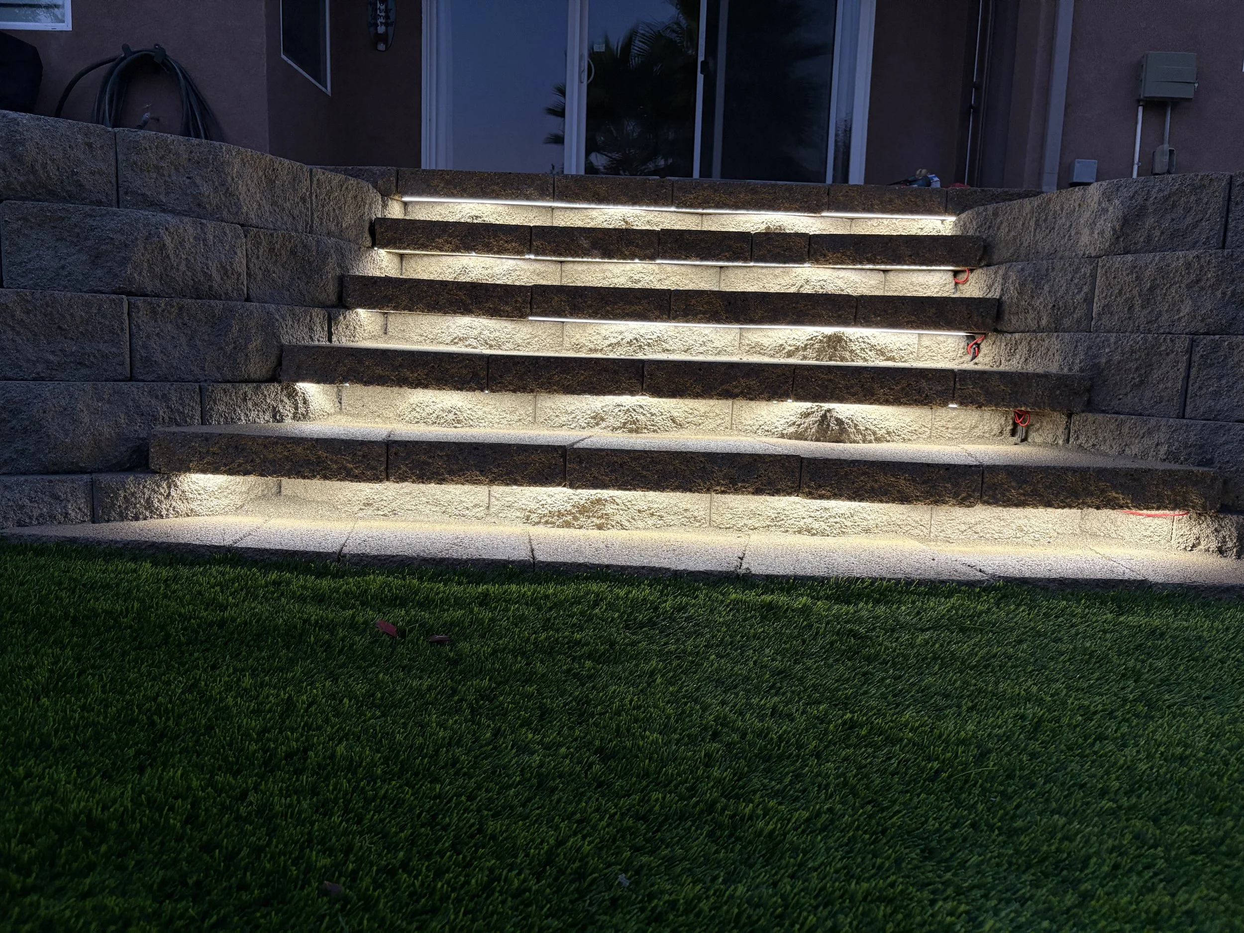 Exterior staircase with illuminated steps leading to a door, flanked by stone walls, at dusk or nighttime. Close-up of natural-color locking stone steps with integrated HD lighting in Canyon Lake, CA.