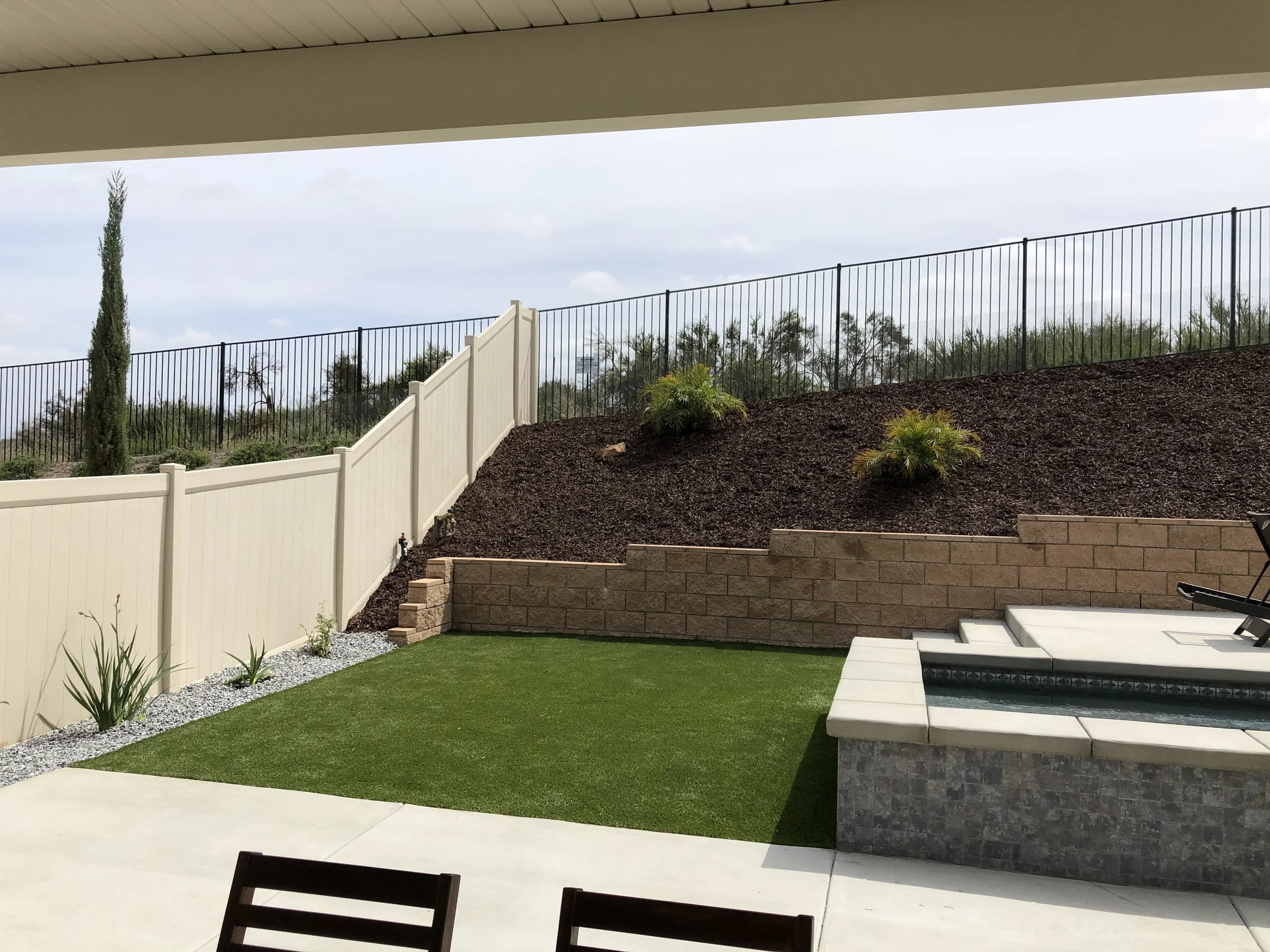 Backyard patio with a small pool, a patch of green artificial grass, and landscaped hillside with soil and plants, enclosed by a white privacy fence and black metal fence.