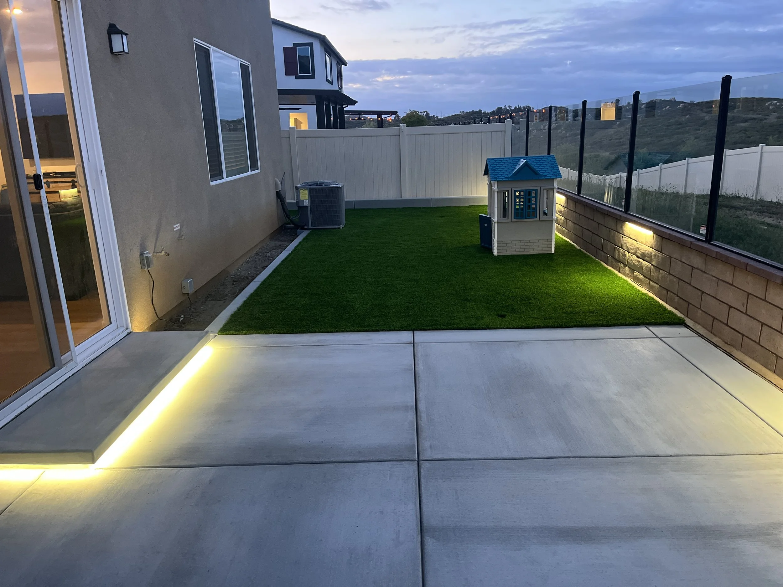 Backyard patio area with artificial grass, small playhouse, and glass railing at sunset.