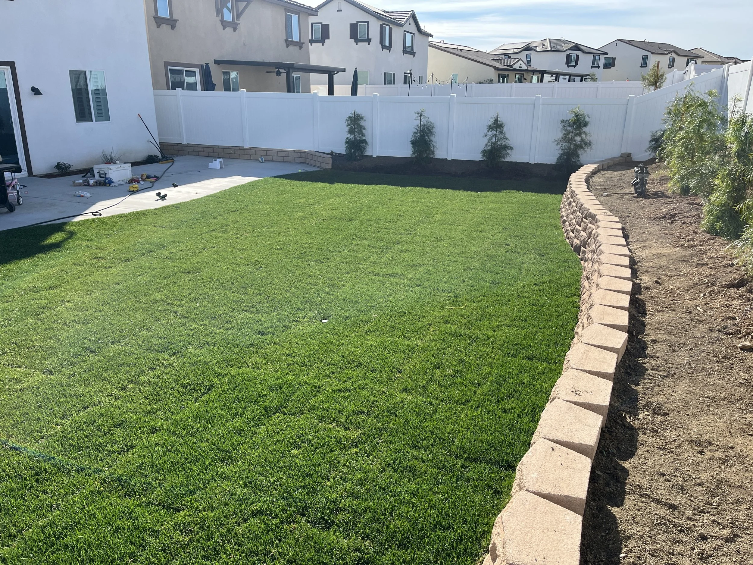 Backyard with a freshly laid green lawn, a white fence, and a stone border on the right side. Construction tools and supplies are scattered near the house.